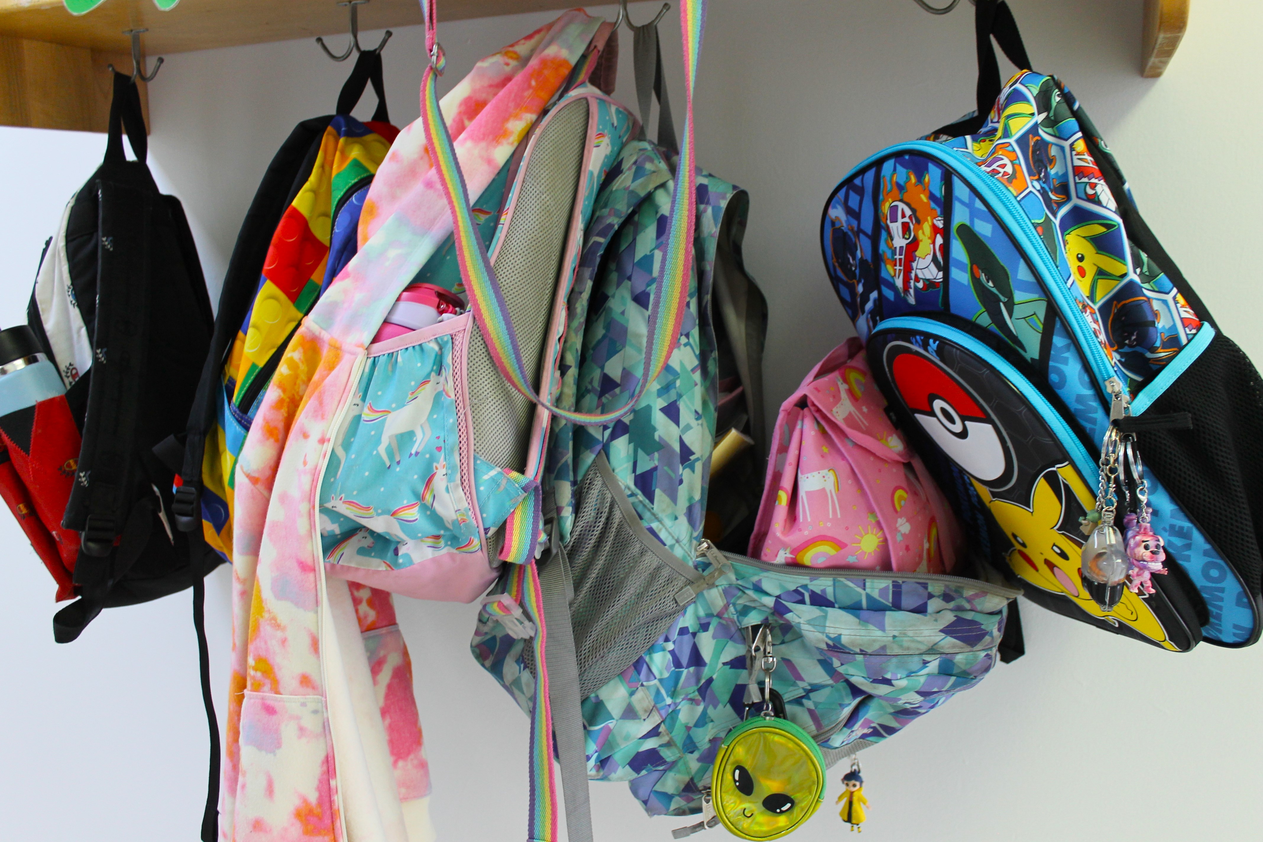 A row of colorful backpacks and coats hanging on wall hooks, including unicorn and Pokémon designs.