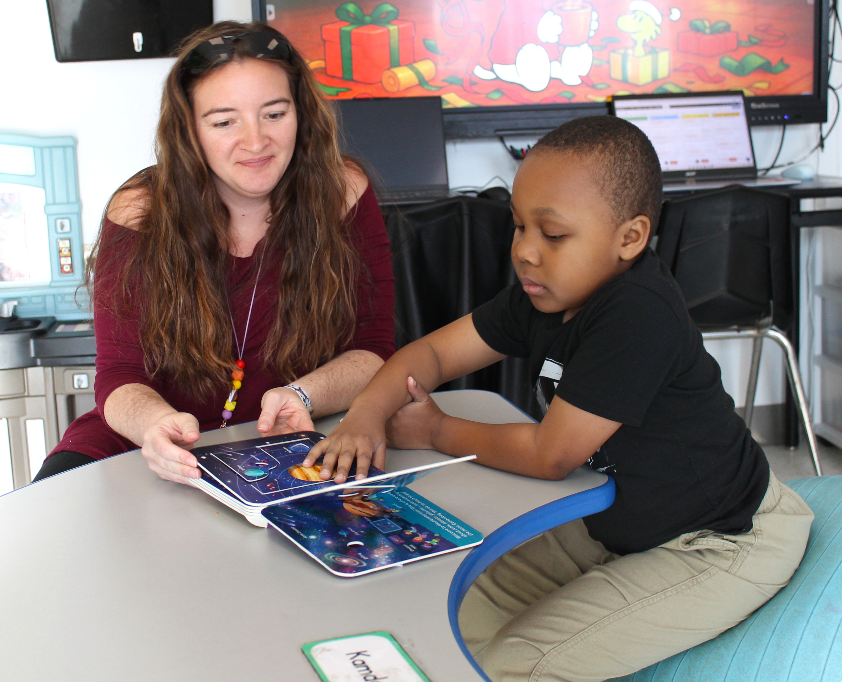 Young boy reading a space-themed book with a woman at a table in a classroom setting.