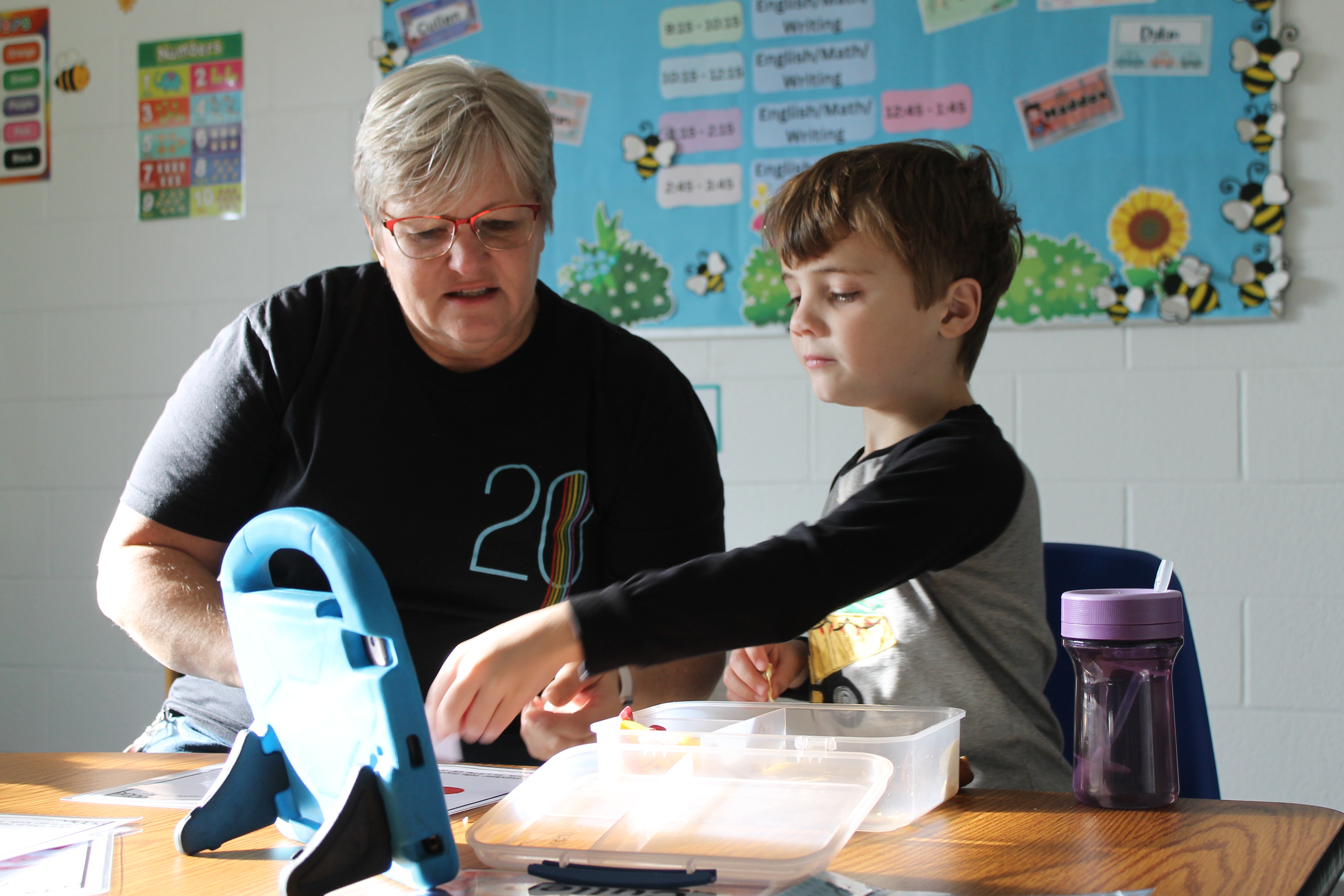 An adult woman wearing glasses and a black shirt assists a young boy with an activity at a table in a classroom.