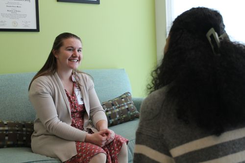 A smiling woman with light brown hair wearing a floral dress and cardigan seated on a couch, talking to another person with curly hair seen from behind.