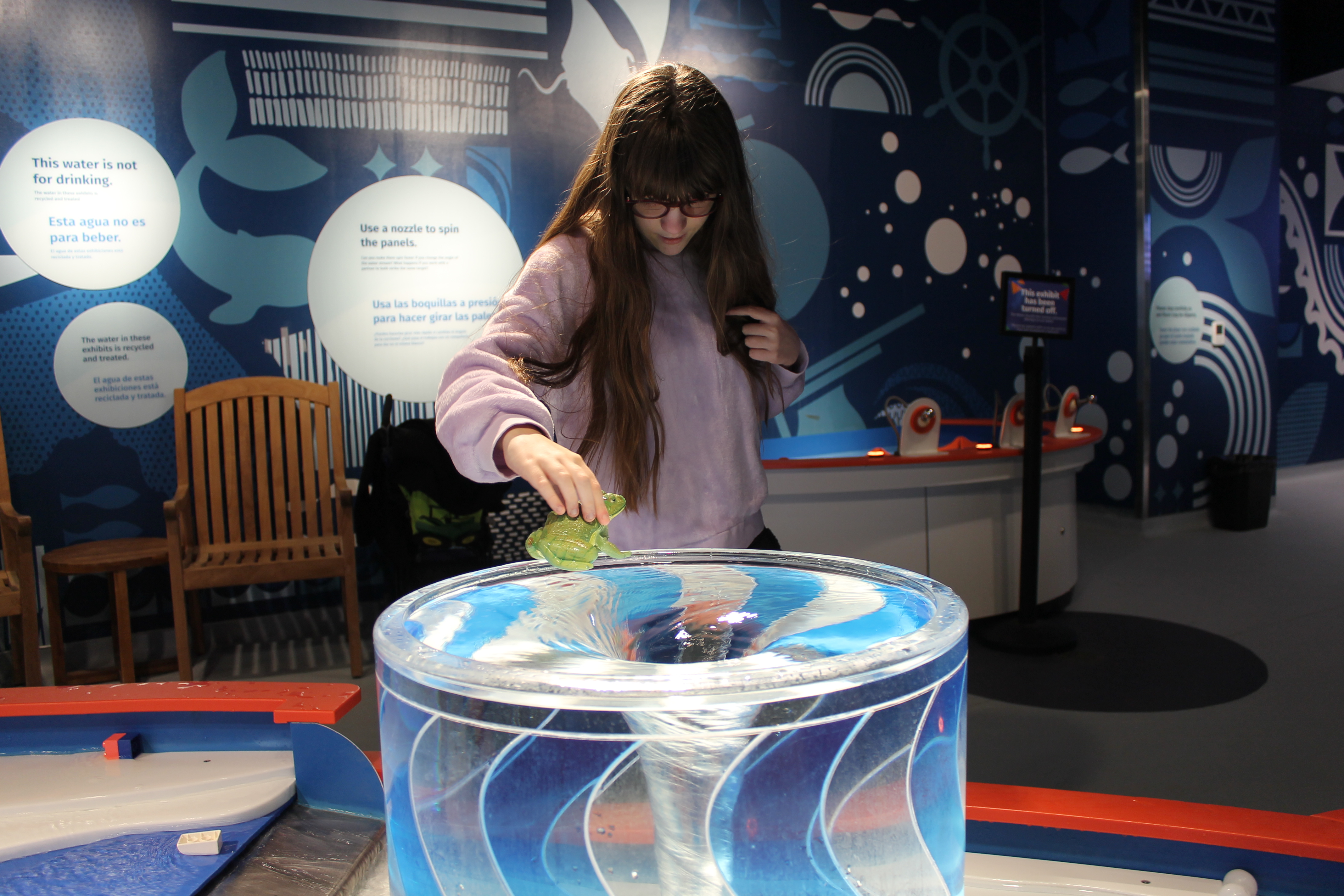 Young girl with long hair and glasses touching a green turtle toy above a cylindrical water vortex exhibit at a science center.