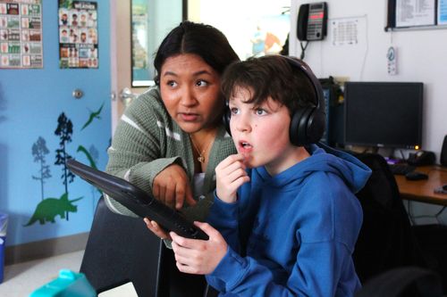 Woman in a green sweater helping a boy in a blue hoodie with headphones using a tablet in a classroom.
