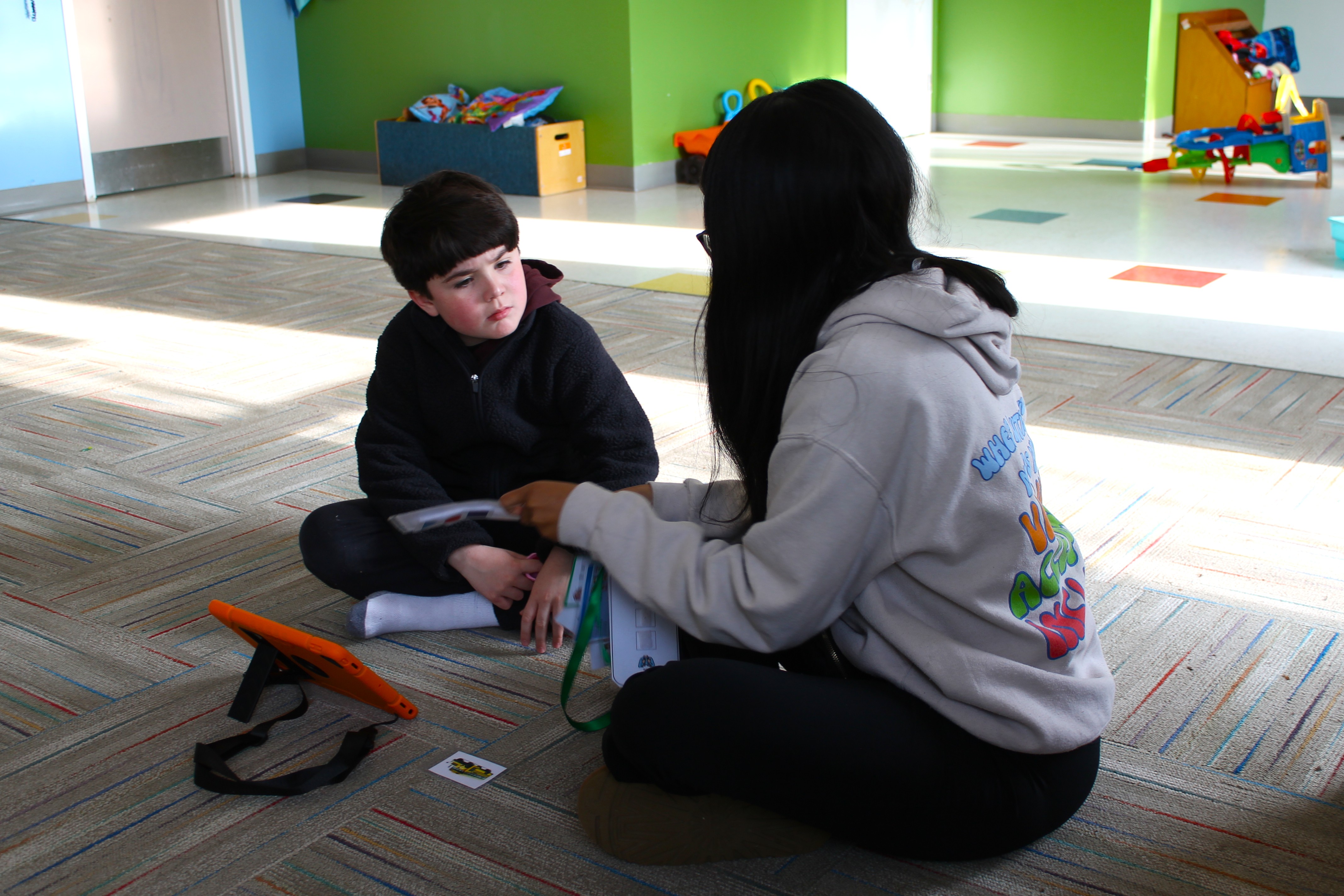 Child sitting on carpeted floor facing an adult who is showing a card, with a tablet in an orange case nearby.