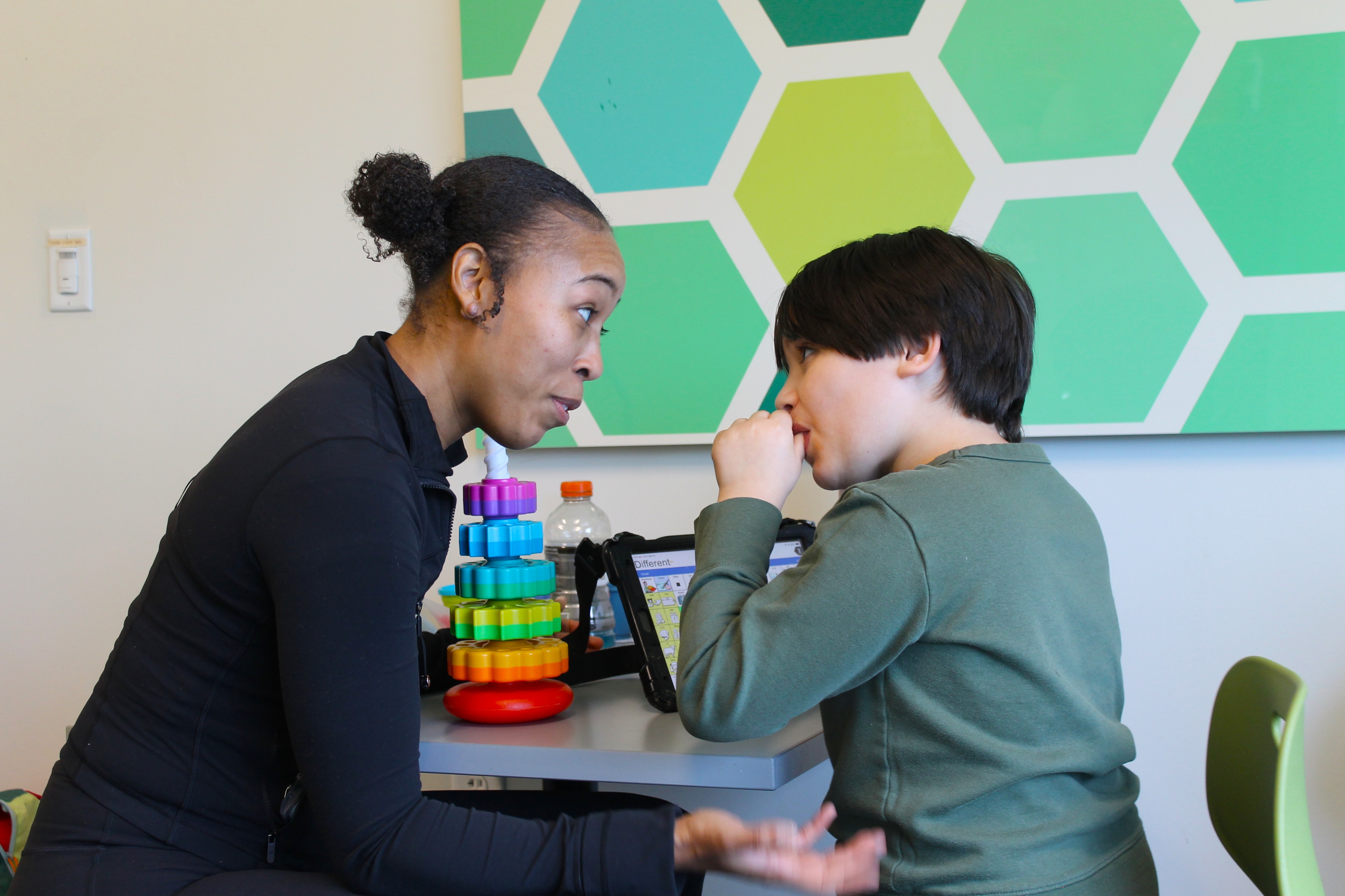 Woman and child engaged in a focused conversation at a table with colorful stackable toys and a tablet device.