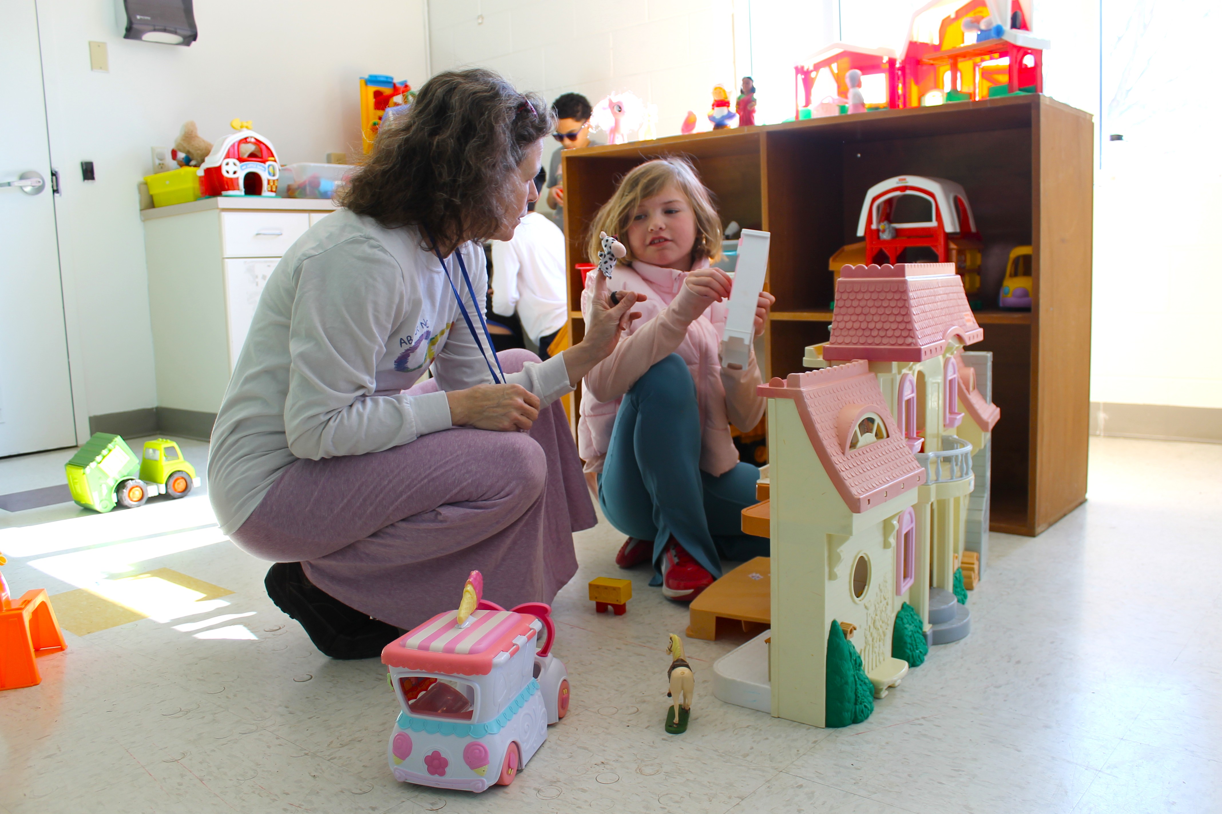 A woman crouching on the floor playing with a young girl near a dollhouse and toys in a well-lit playroom.