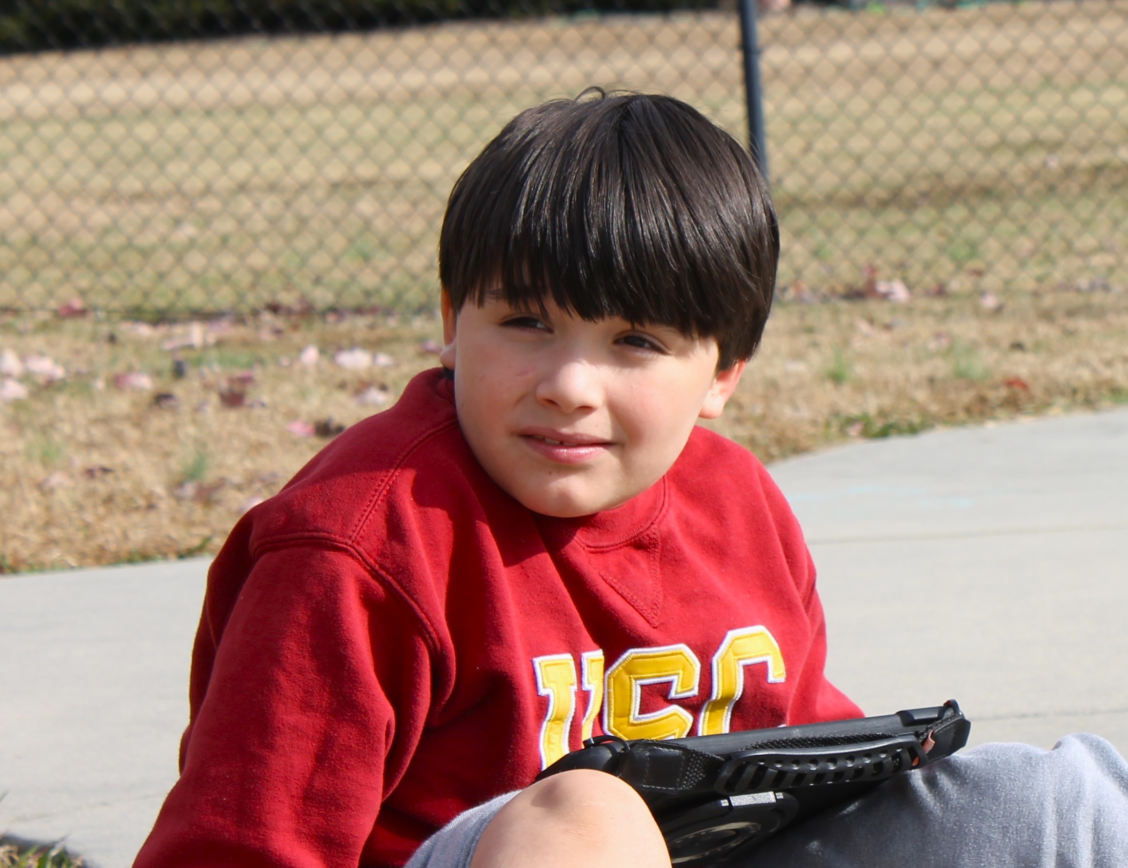 Young boy in a red USC sweatshirt sitting outdoors on a concrete surface with a tablet on his lap.