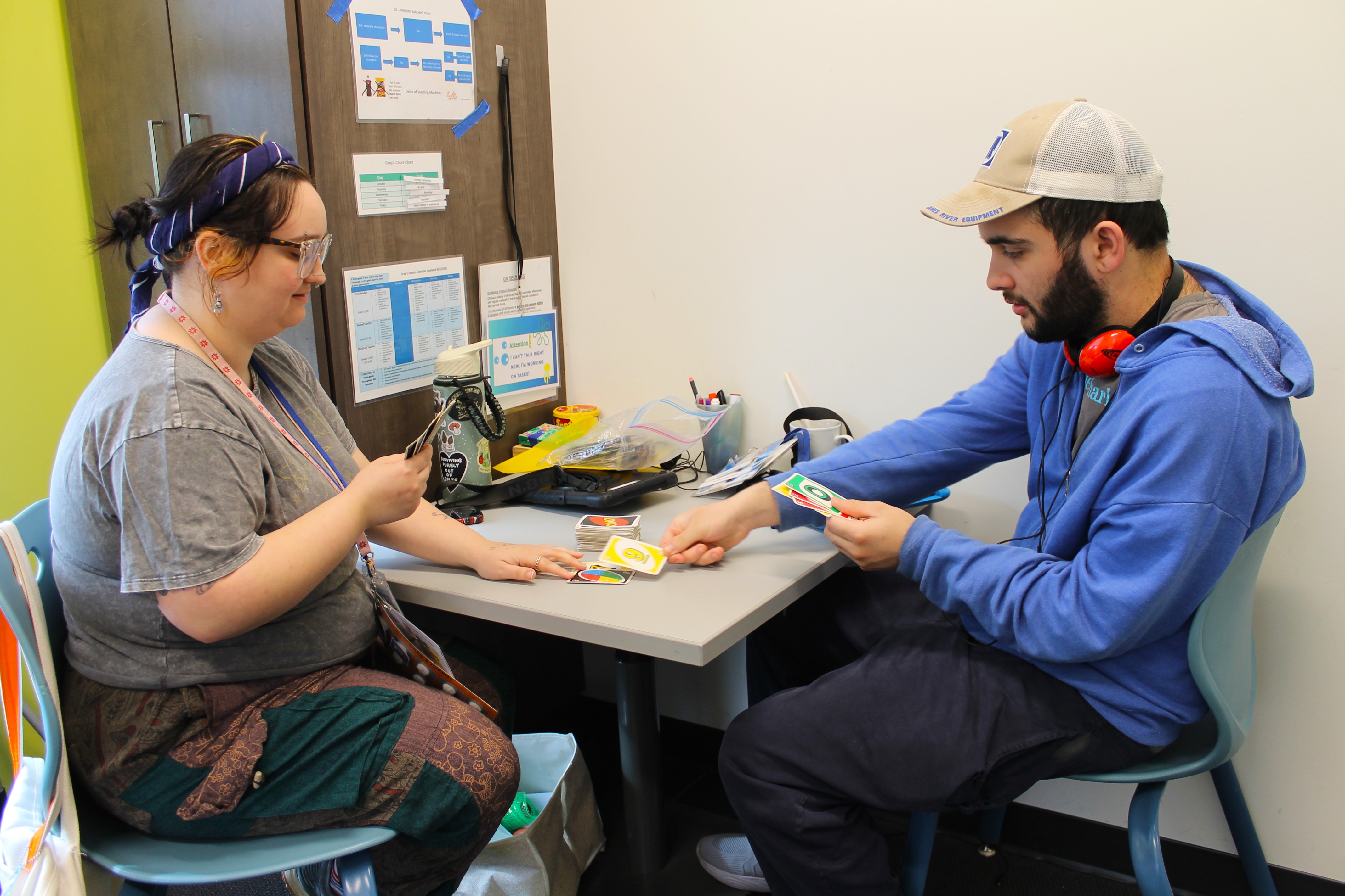 Two people sitting at a table playing the card game Uno, with one person placing a yellow card down.
