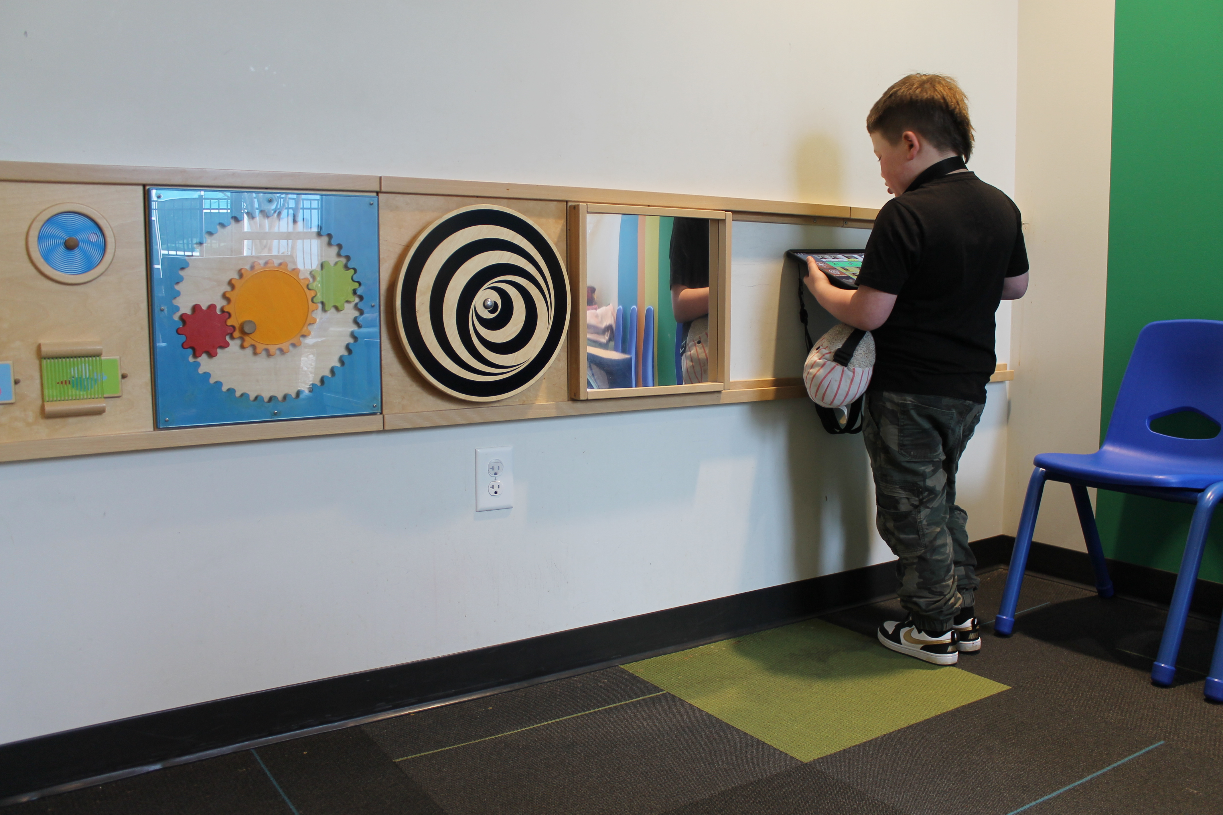 Child standing and interacting with a wall-mounted activity panel featuring colorful gears, a spiral pattern, and a small mirror, next to a blue chair.