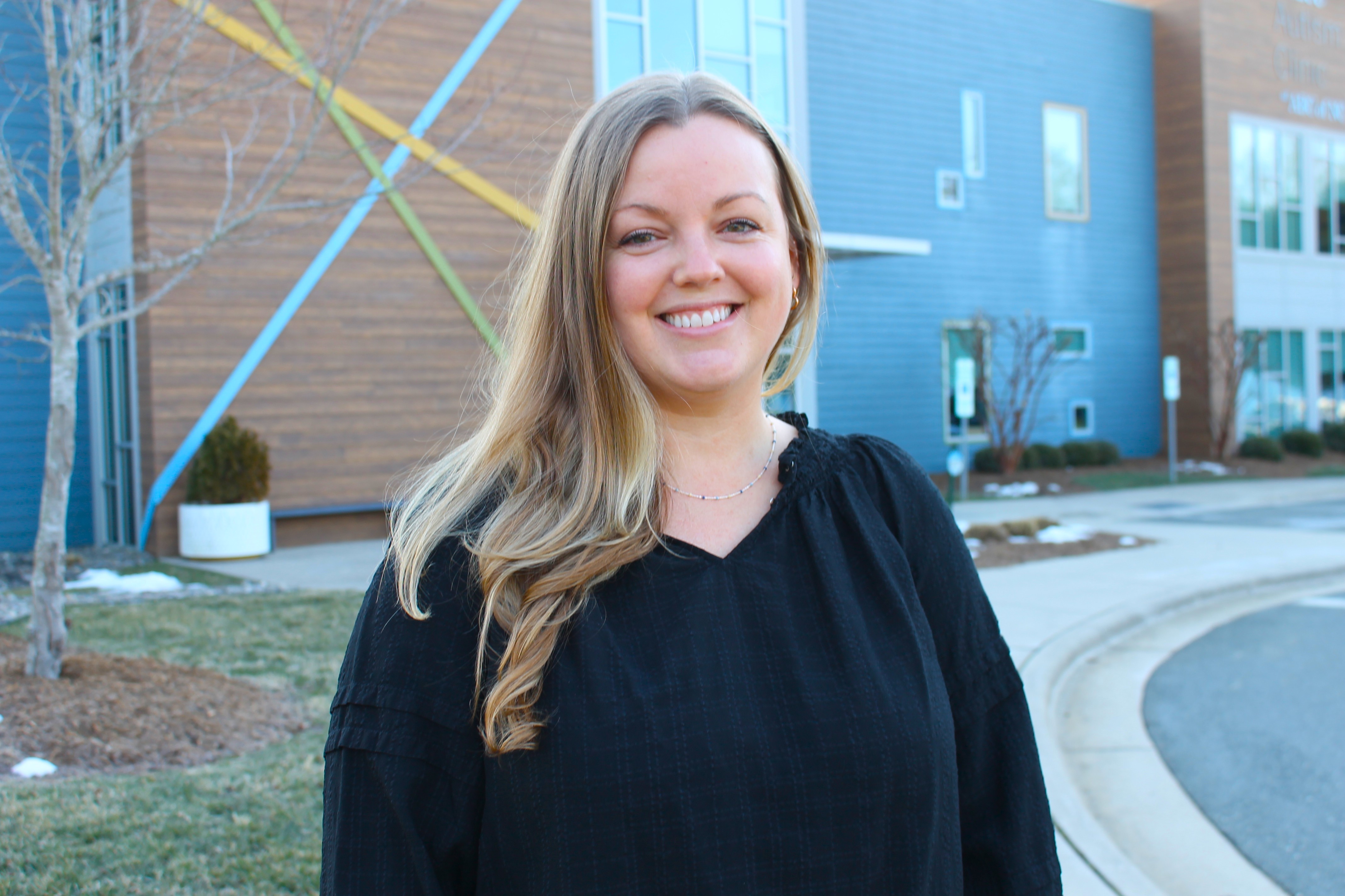 Smiling woman with long blonde hair wearing a black top standing outdoors near a modern building.
