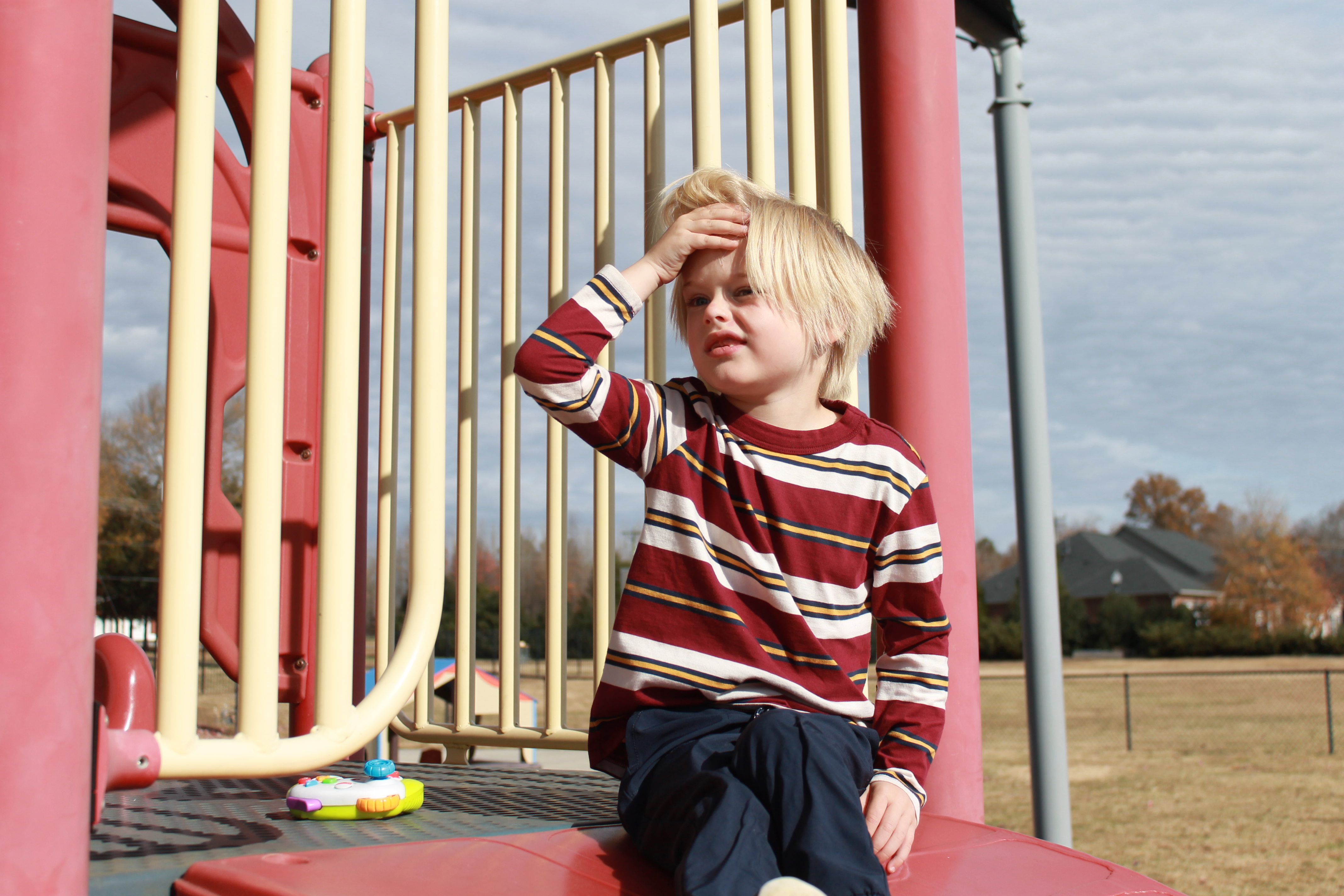 Young boy with blonde hair sitting on a playground structure, holding his forehead with one hand and looking to the side.