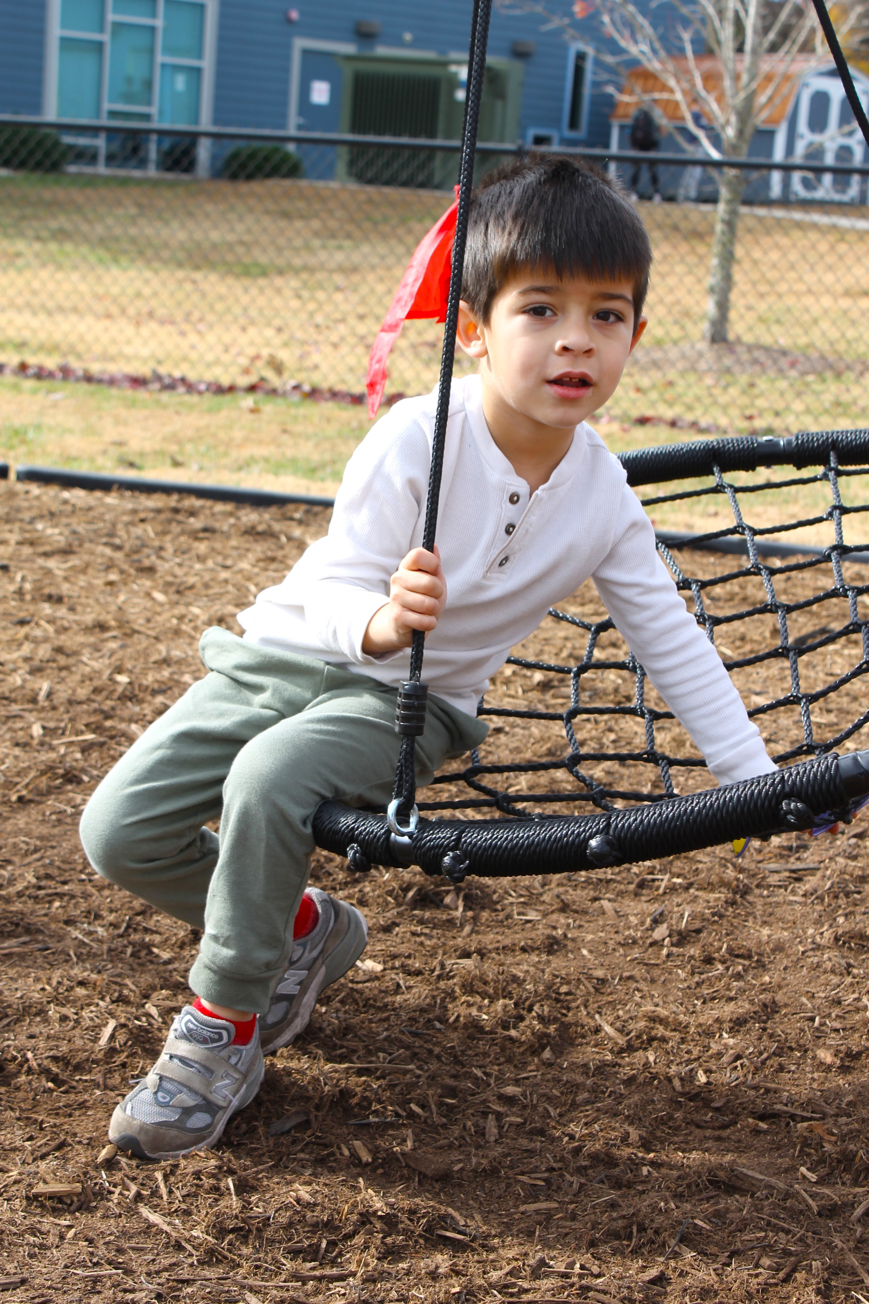 Young boy in white shirt and green pants sitting on a black net swing at a playground.