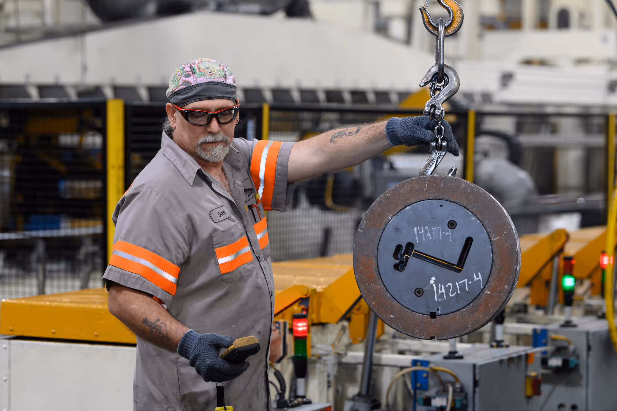 Factory worker wearing safety glasses and gloves operating a crane hook lifting a large metal part in an industrial setting.