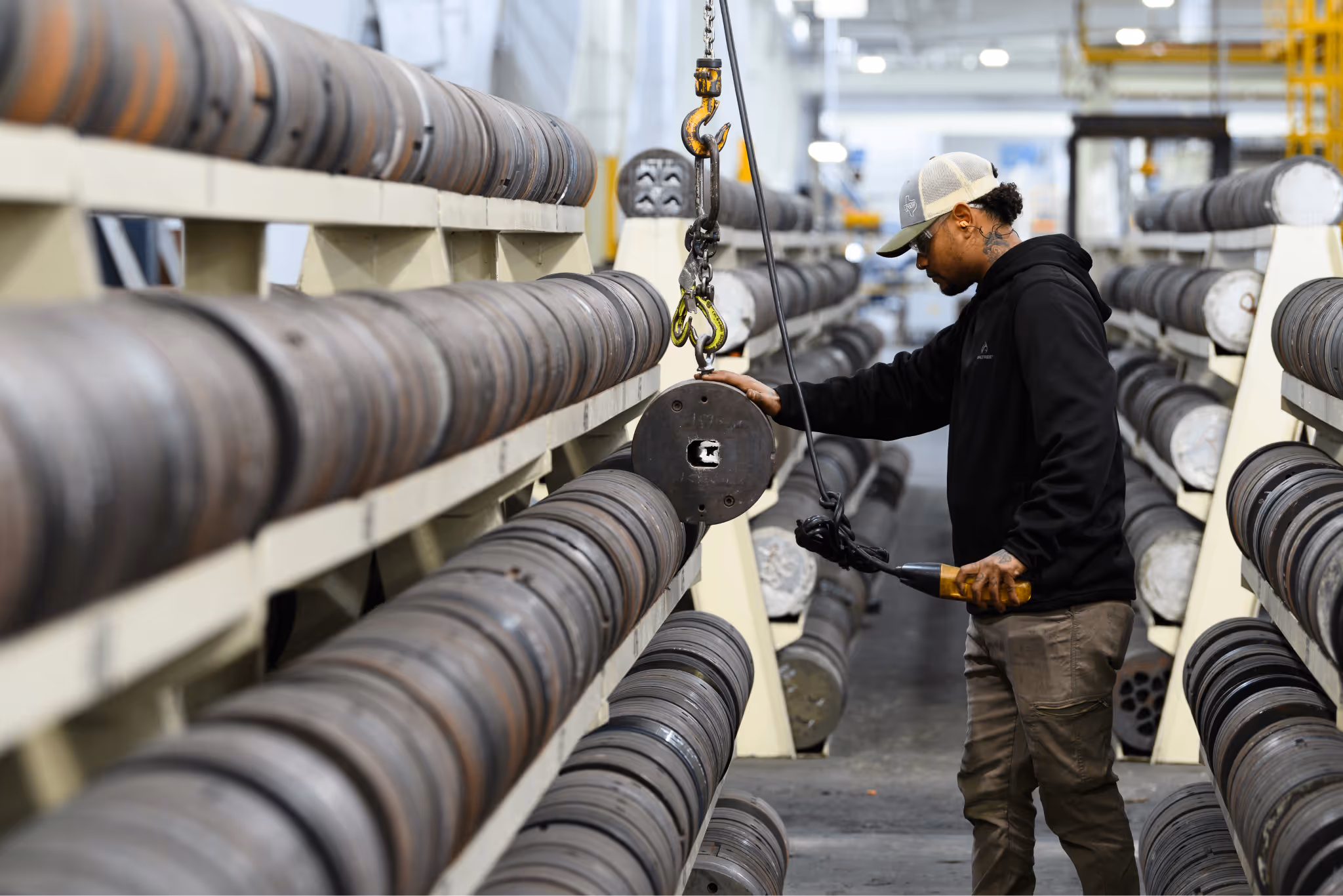 Worker operating a hoist hook to lift a metal spool in an industrial storage area filled with rows of metal spools.