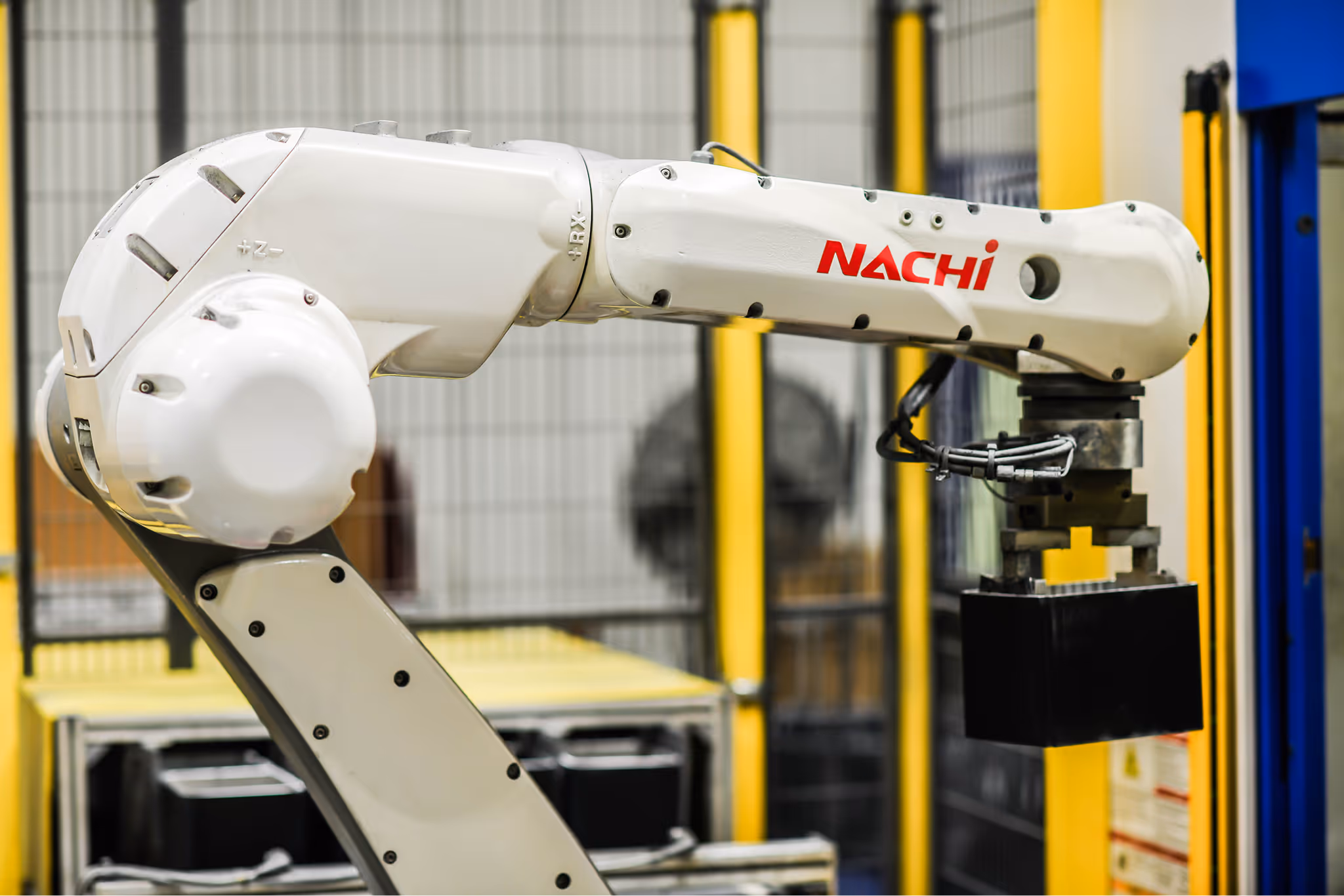 Close-up of a white industrial robotic arm labeled Nachi holding a black object inside a factory setting.