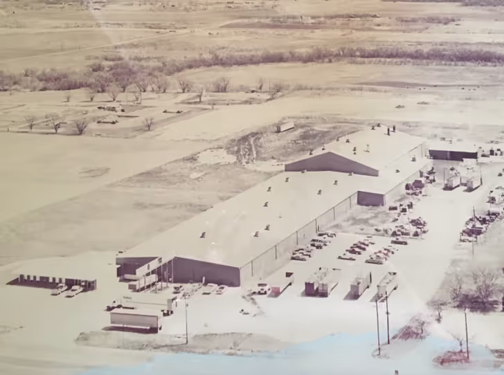 Aerial view of a large industrial warehouse complex surrounded by parked vehicles and open land.