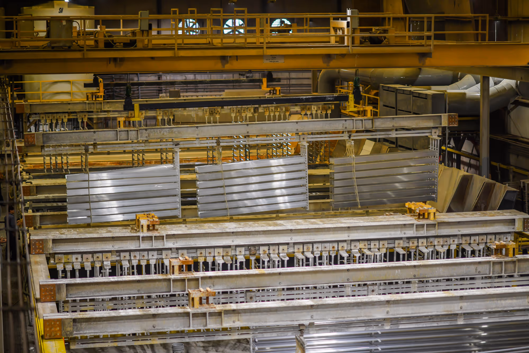 Metal sheets suspended by hooks in an industrial anodizing facility with yellow railings and machinery.