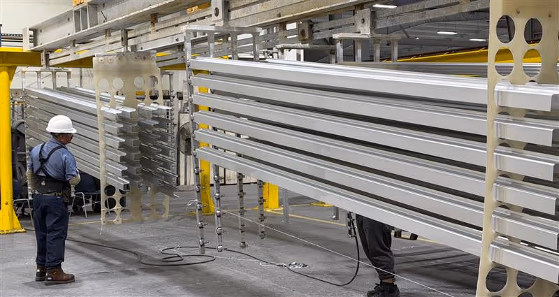 Two workers in safety gear inspecting hanging long aluminum extrusions in an industrial facility.