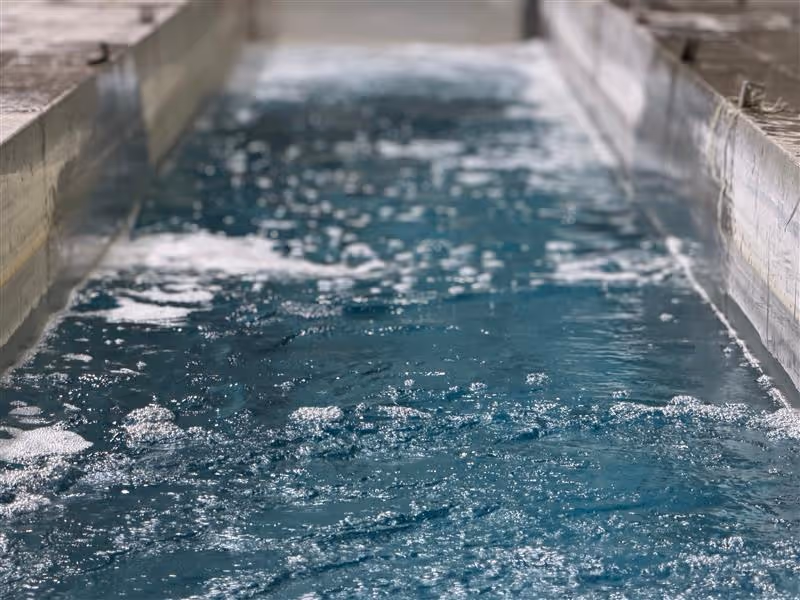 Blue water flowing rapidly through a narrow concrete channel with bubbles and ripples, showing the anodize process.