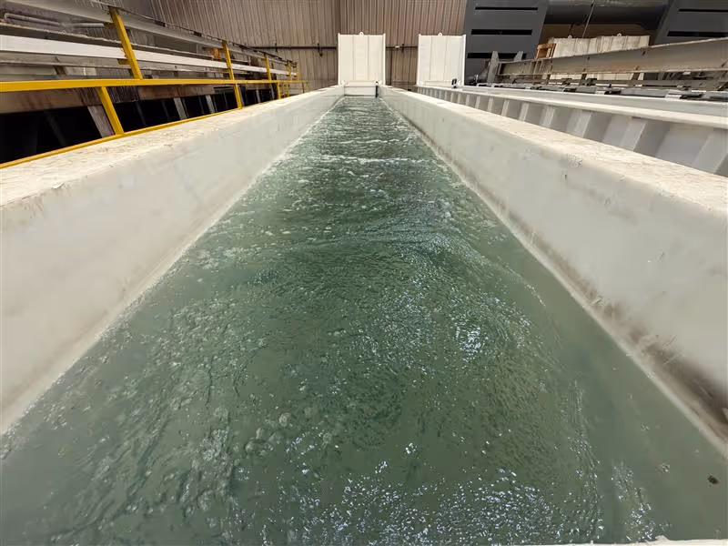 Water flowing vigorously through a long, white industrial channel inside an aluminum anodize facility.