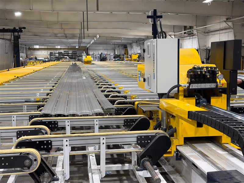 Industrial conveyor system transporting long metal beams inside a factory with yellow machinery.