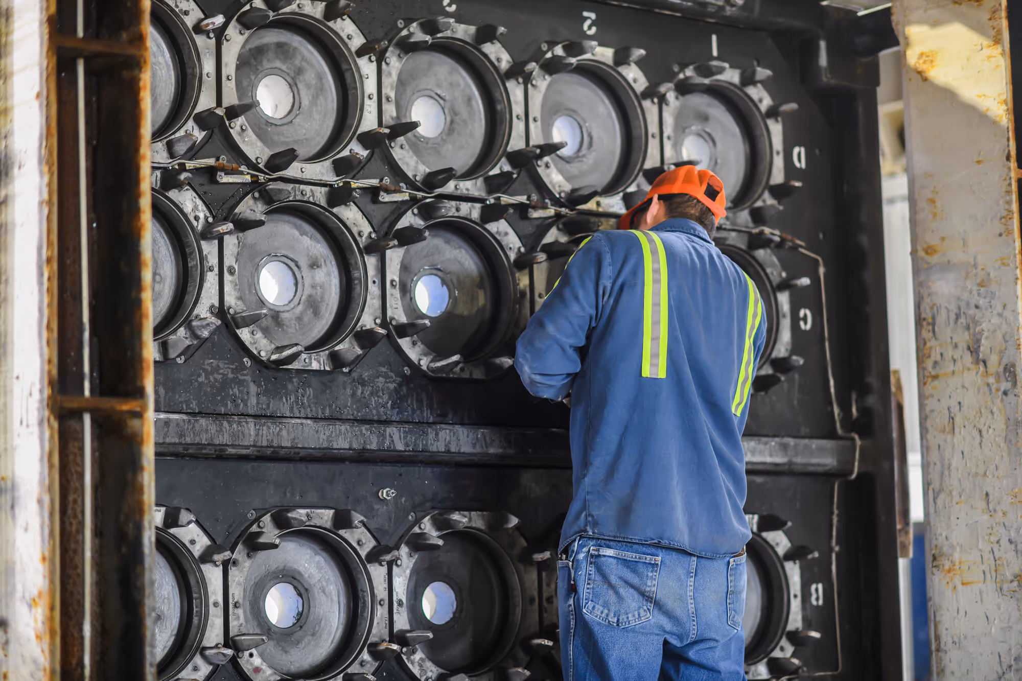 Worker in blue jacket and orange hard hat inspecting large industrial machinery with multiple circular metal components.