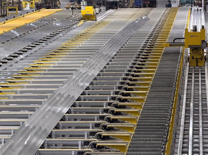 Industrial manufacturing line with automated yellow machinery handling rows of long metal beams.