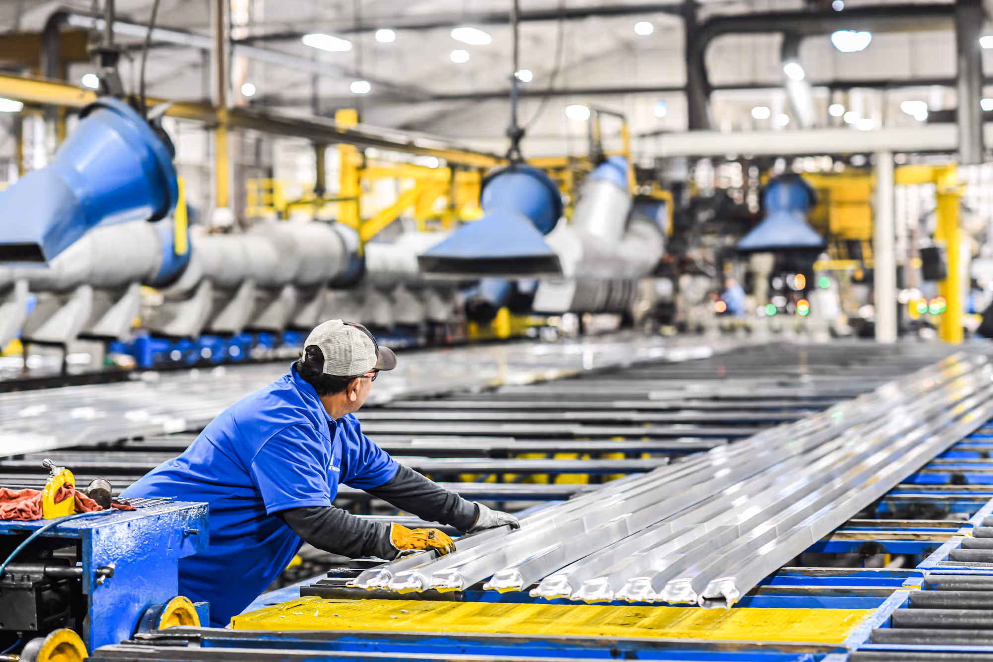Worker in blue shirt and cap handling long metal profiles on a factory production line.