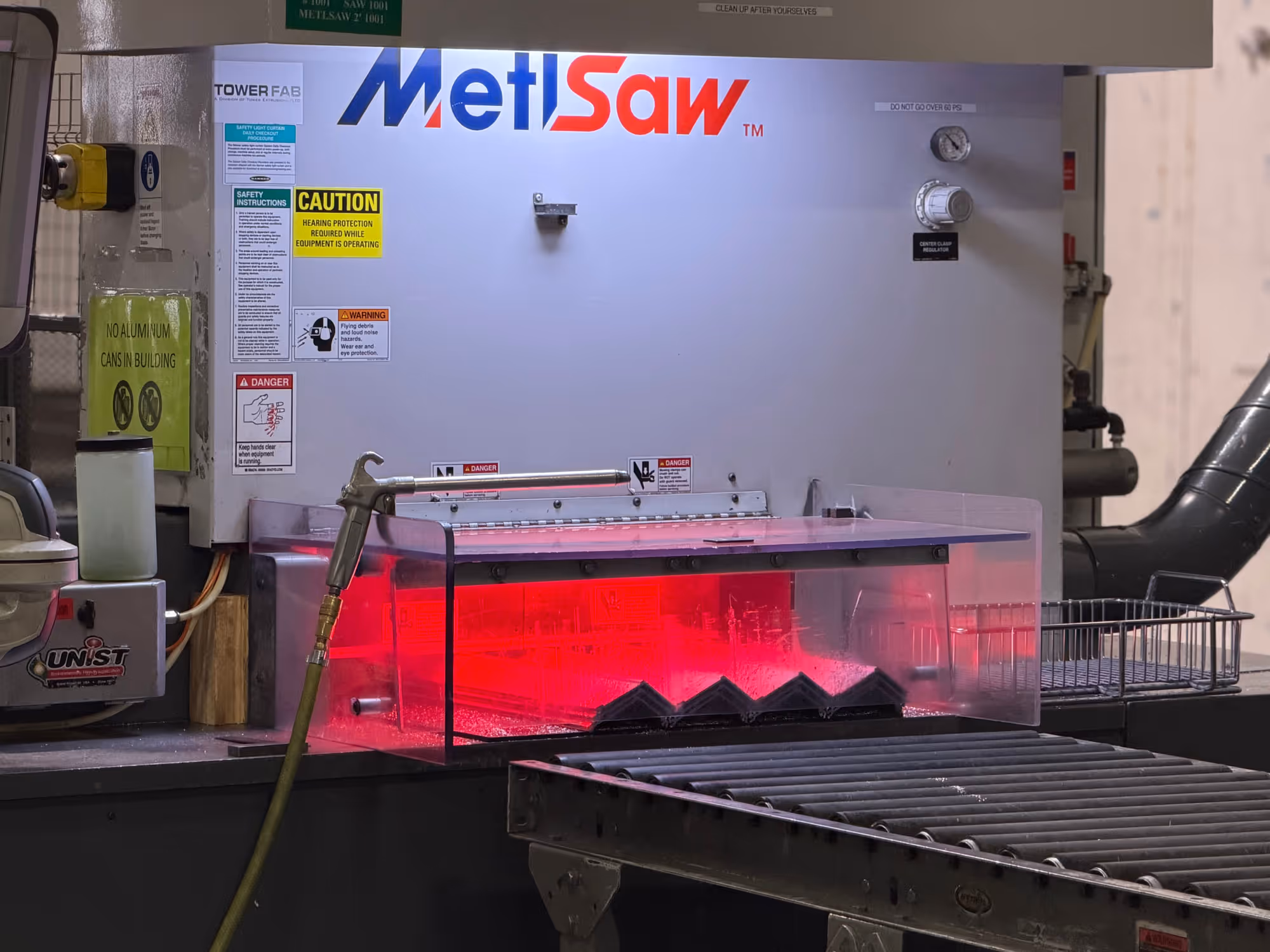 Industrial MetlSaw metal cutting machine with safety warnings and roller conveyor in a workshop.