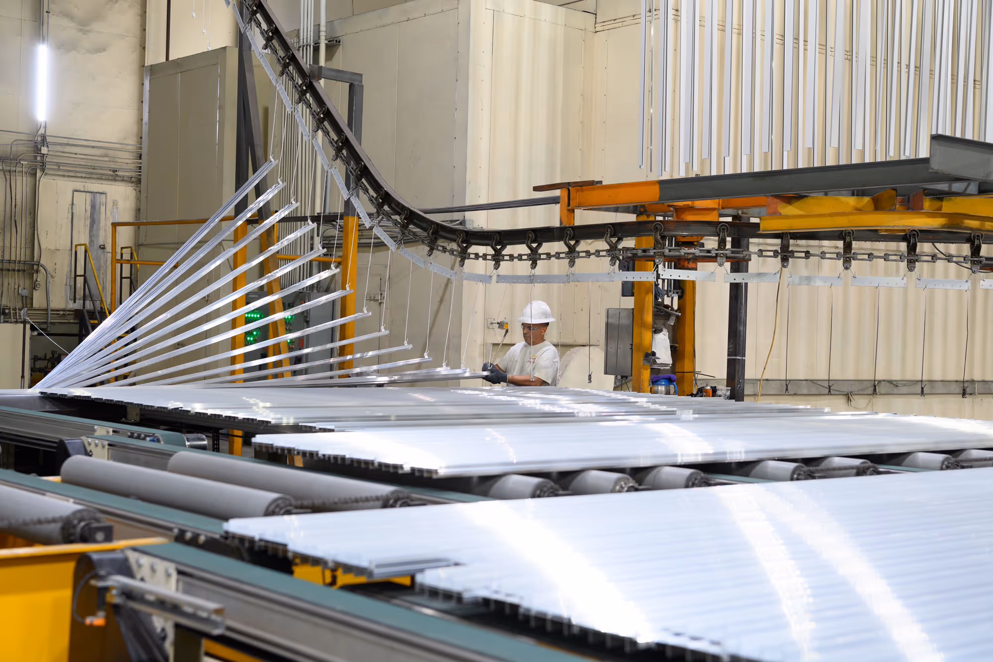Worker wearing a hard hat inspecting metal sheets on a conveyor system in an industrial facility.
