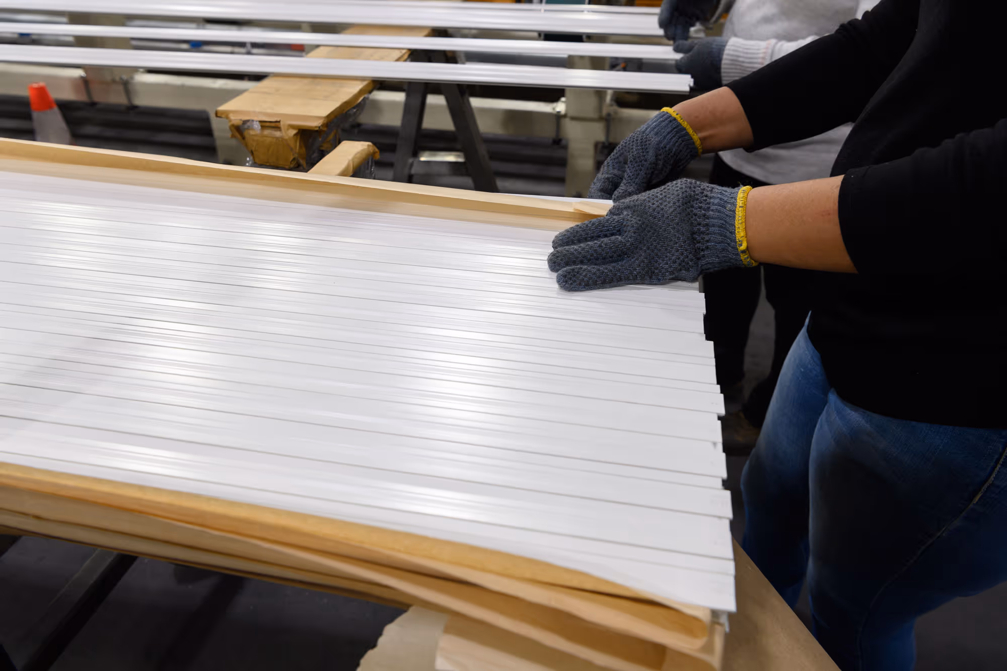 Person wearing gray gloves assembling white slatted panels on a wooden frame in a workshop.