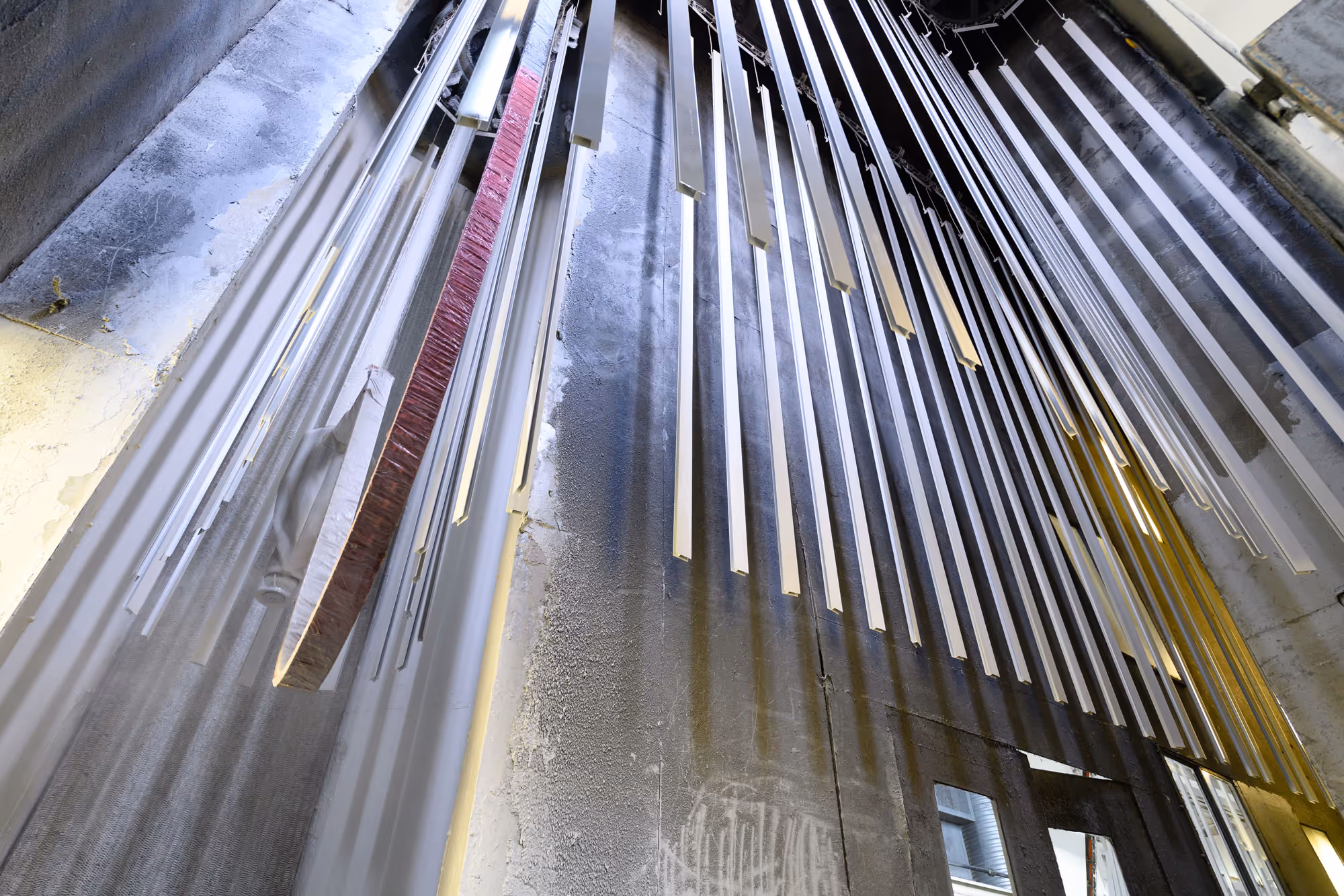 Long, narrow metal chimes hanging vertically in an industrial space with a textured wall background.