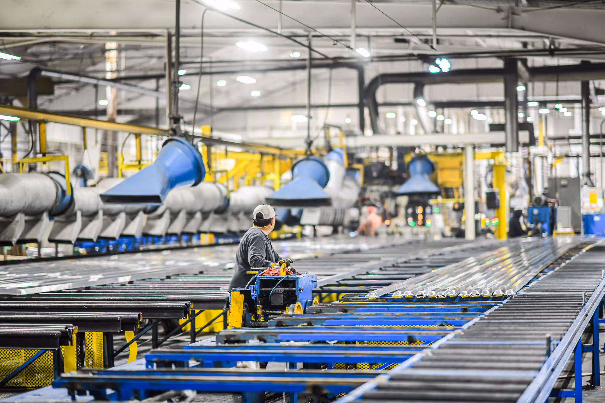 Worker in a production facility operating machinery on a conveyor belt with large ventilation ducts overhead.