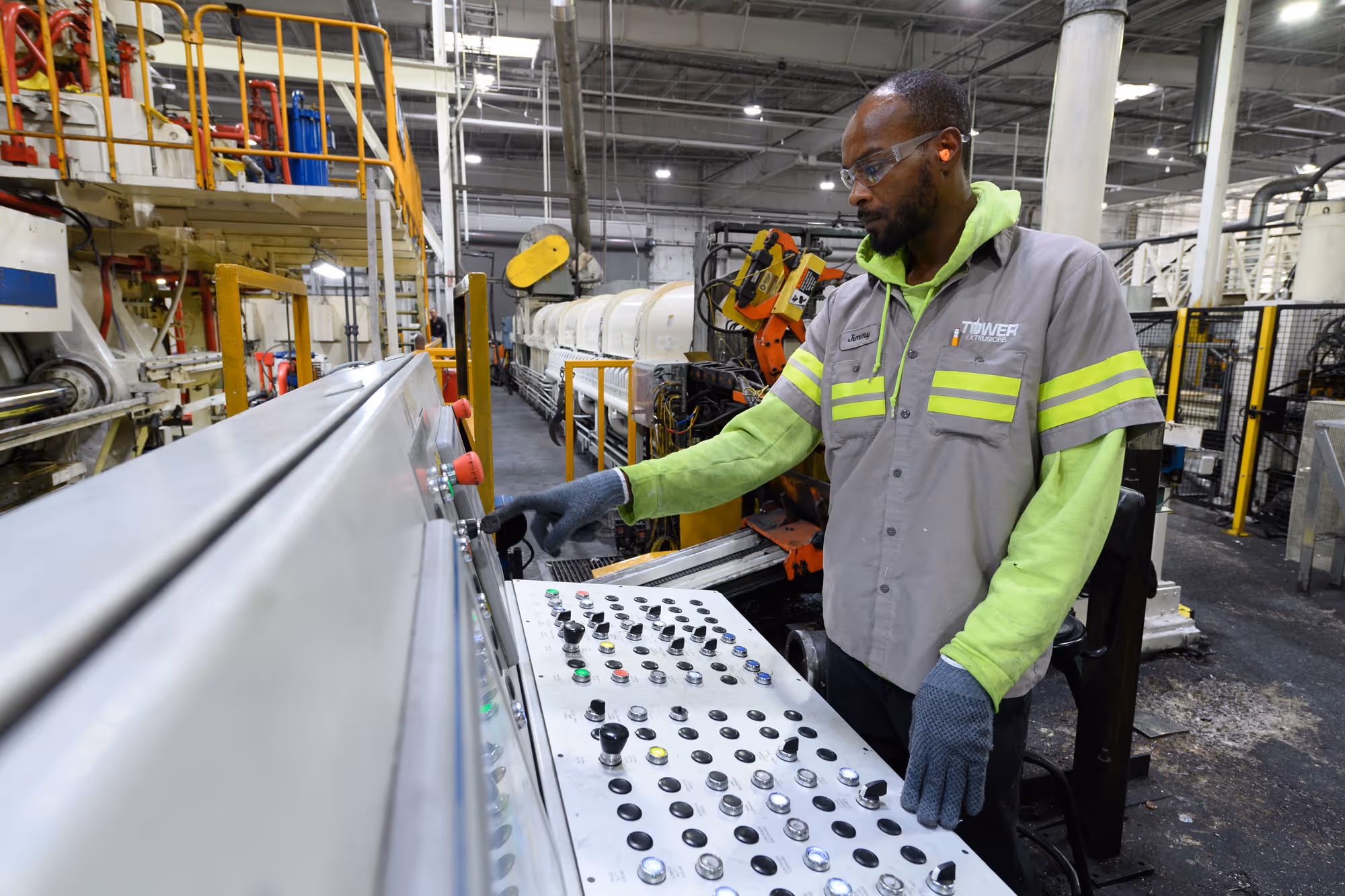 Factory worker wearing safety glasses and gloves operating a large industrial control panel with numerous buttons and switches.