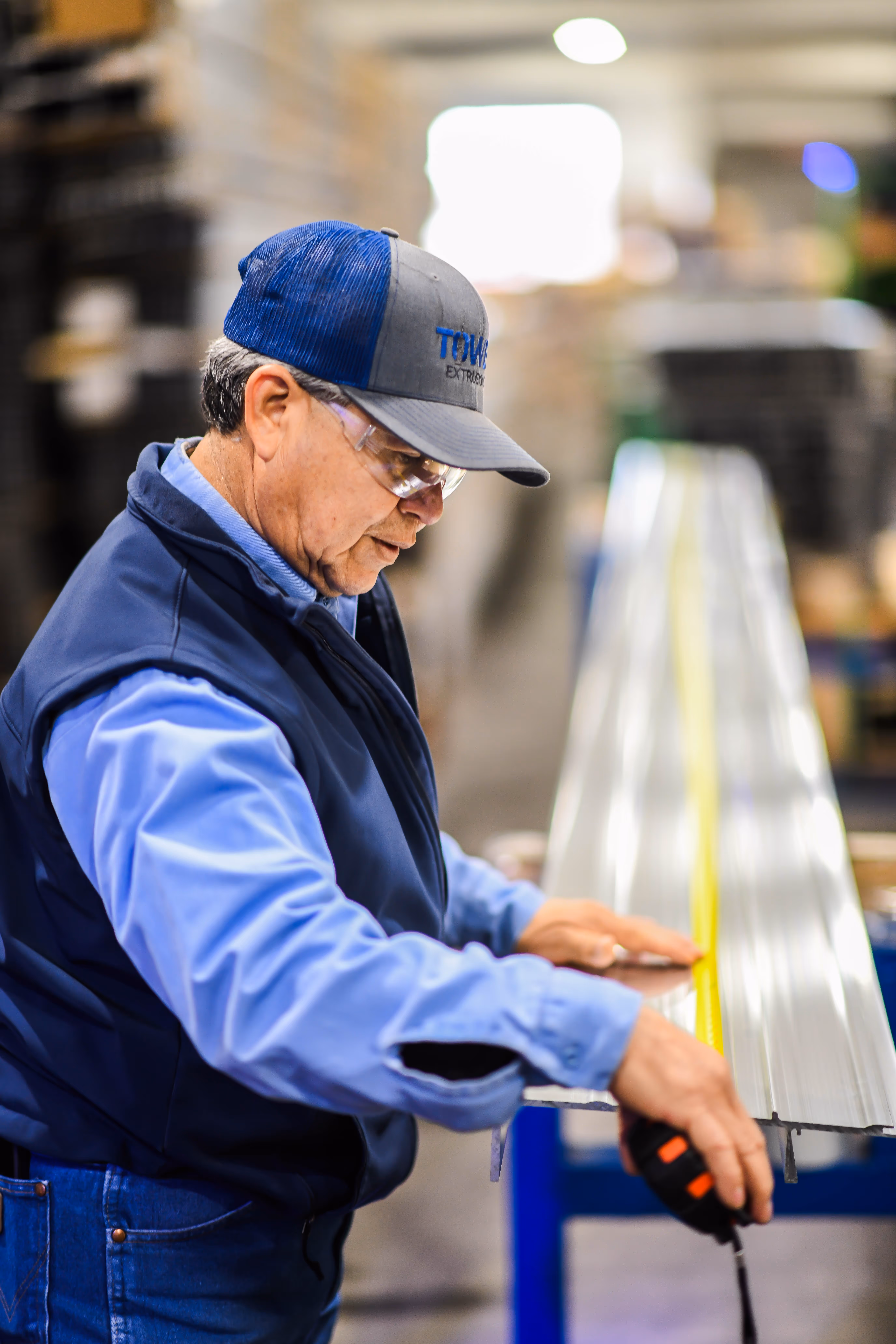 Older man wearing safety glasses and a cap measures a long metal piece with a tape measure in a workshop.