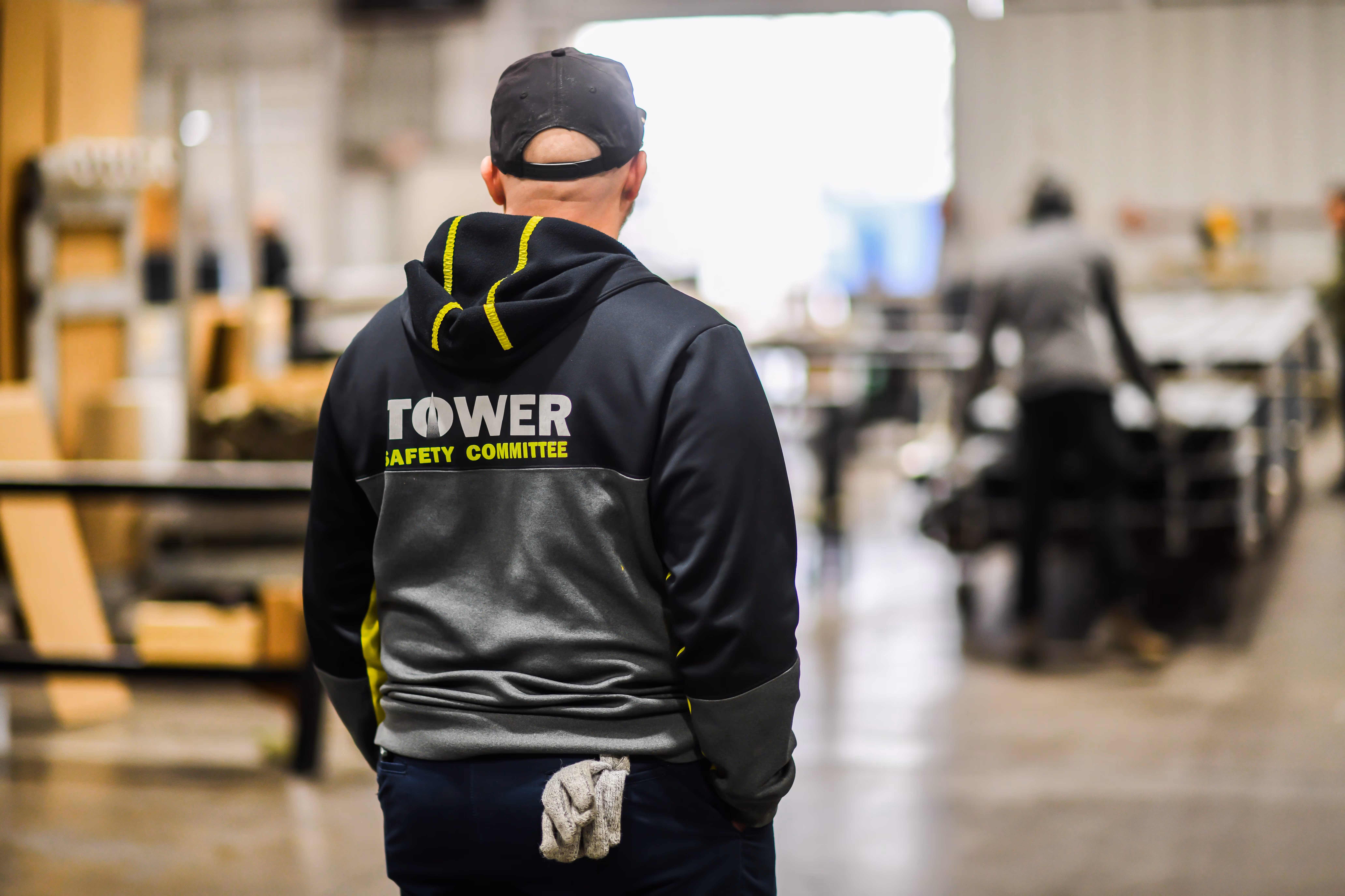 Worker wearing a black and gray hoodie with 'TOWER SAFETY COMMITTEE' on the back inside a warehouse.