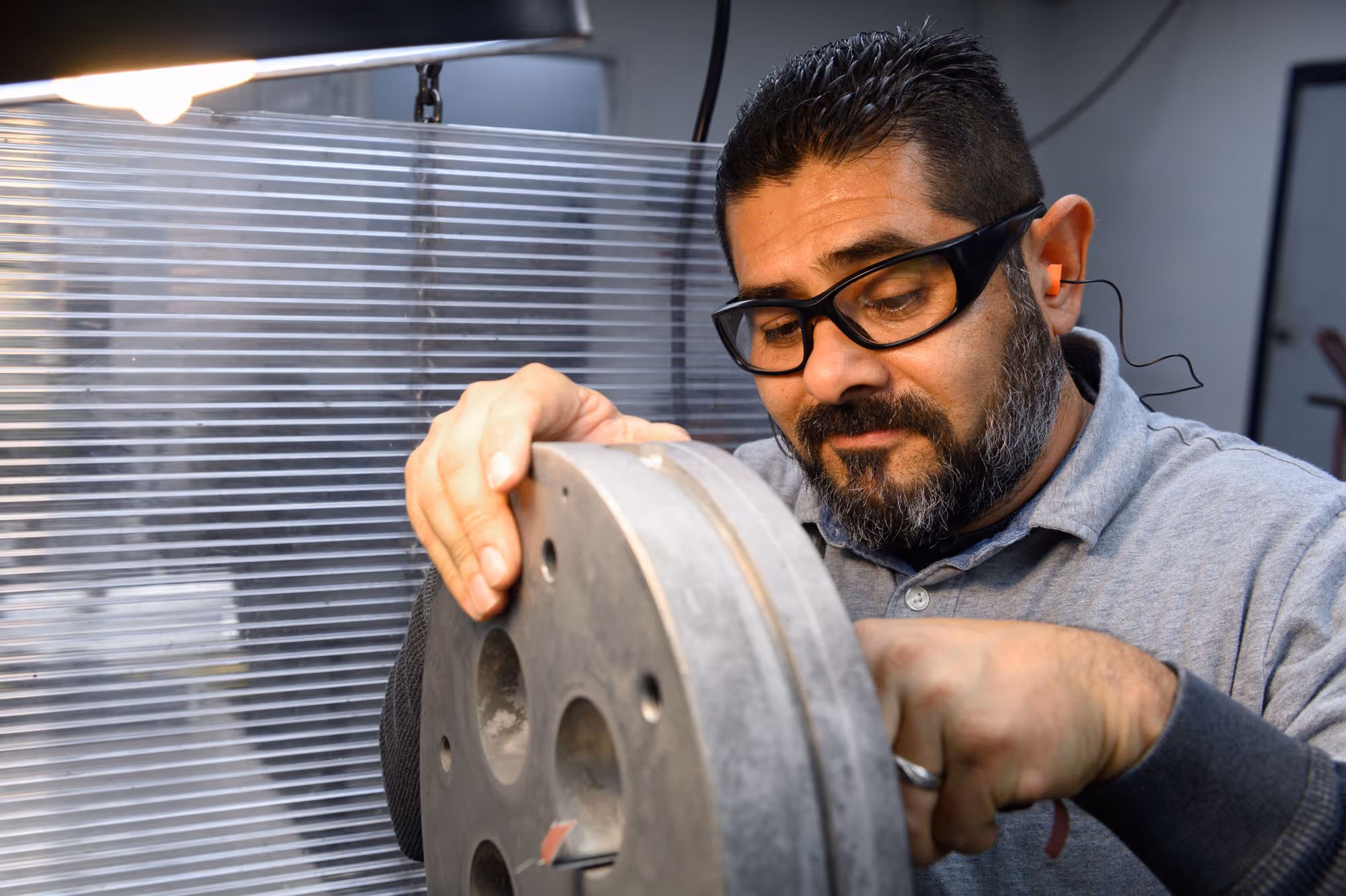 Man wearing safety glasses and ear protection working on a metal wheel or pulley in a workshop.