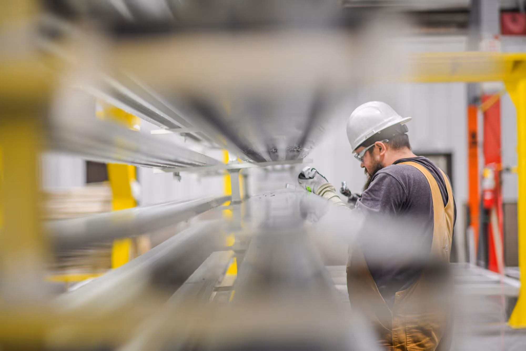 Worker in a white hard hat and brown overalls inspecting metal beams in an industrial warehouse.