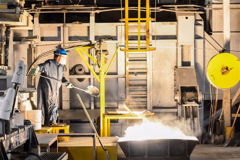 Worker in protective gear handling molten metal in an industrial foundry.