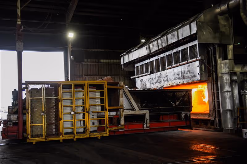 Industrial furnace with metal structure on a conveyor inside a factory with glowing orange heat visible inside the furnace chamber.