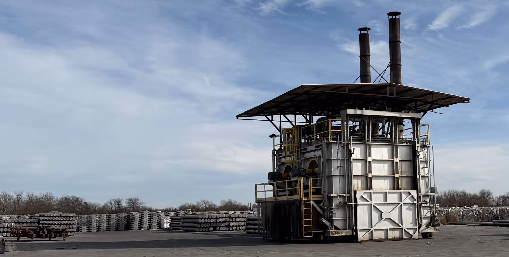 Industrial metal furnace with two chimneys under a roof, surrounded by stacks of cylindrical metal rods outdoors.