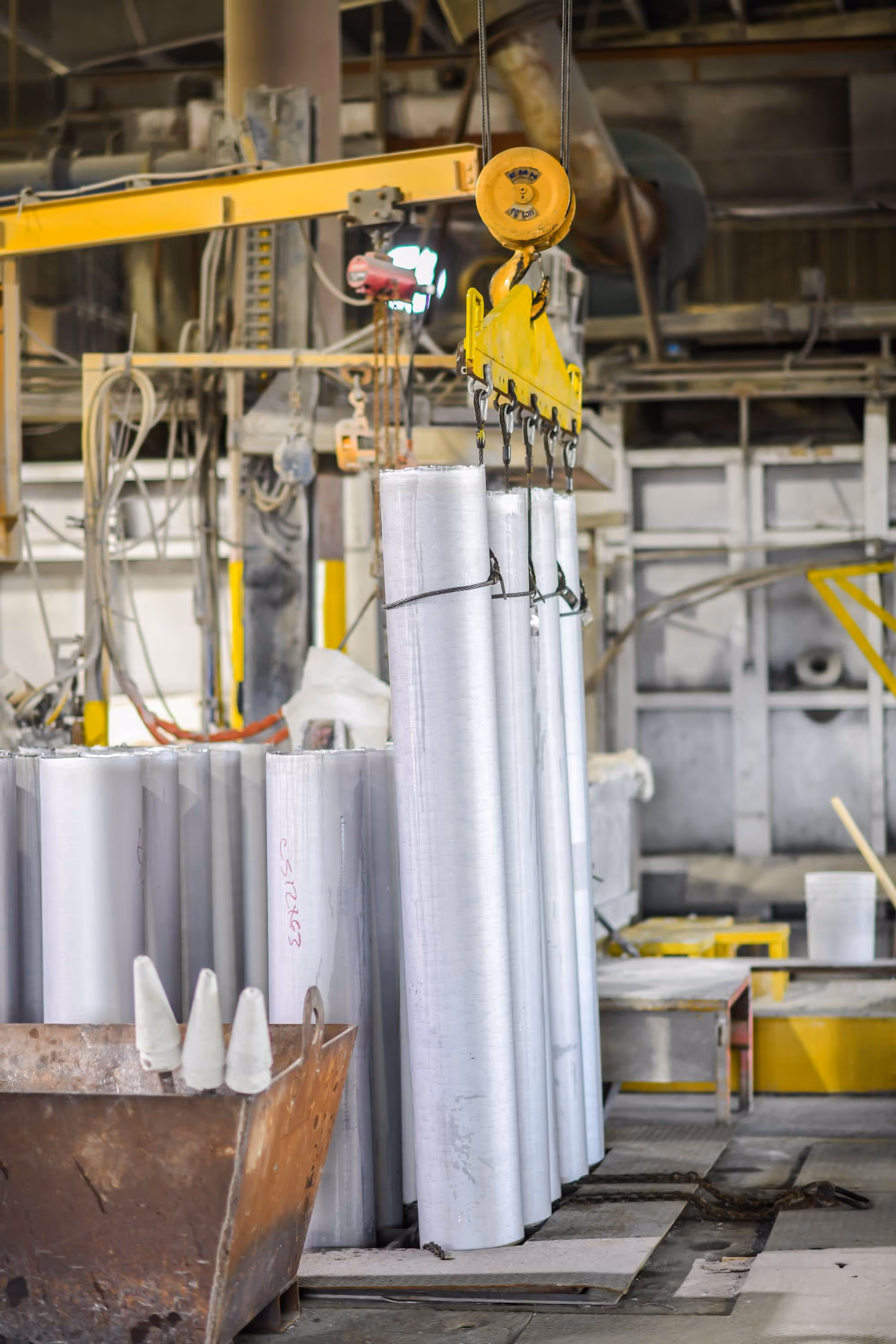 Metal pipes being lifted by a yellow industrial crane hook inside a factory.