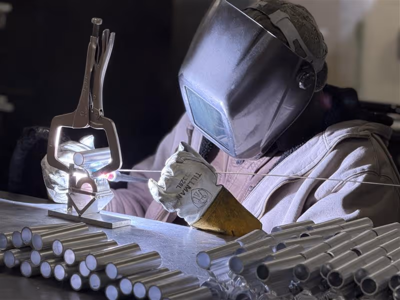 Welder in protective gear welding metal rods on a workbench with several metal rods nearby.
