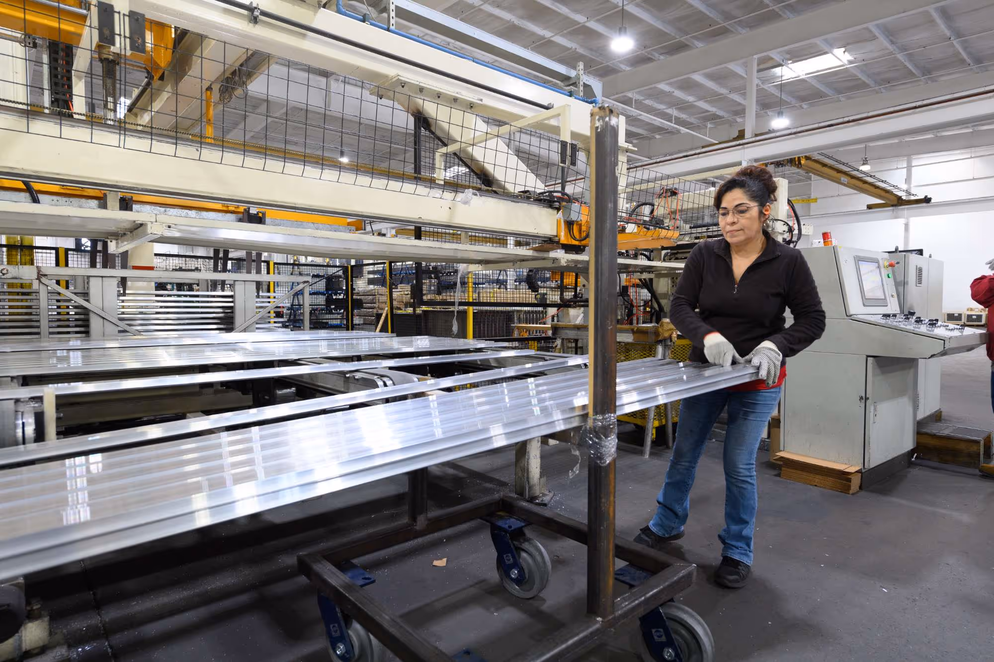 Woman in safety gloves working with metal sheets in an industrial manufacturing facility with machinery and control panels.