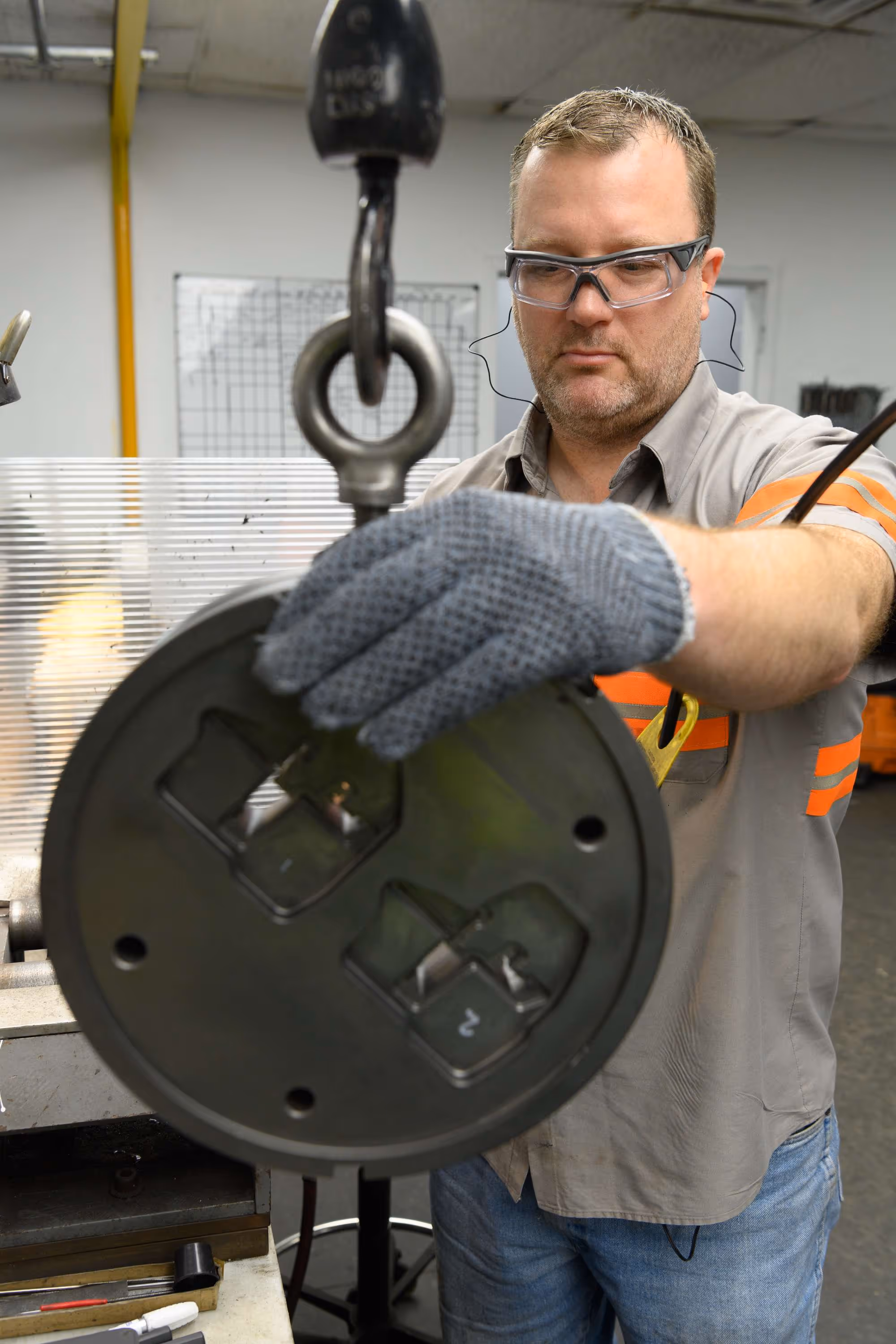 Man wearing safety glasses and gloves operates a hoisting device to lift a heavy circular metal object in an industrial workshop.