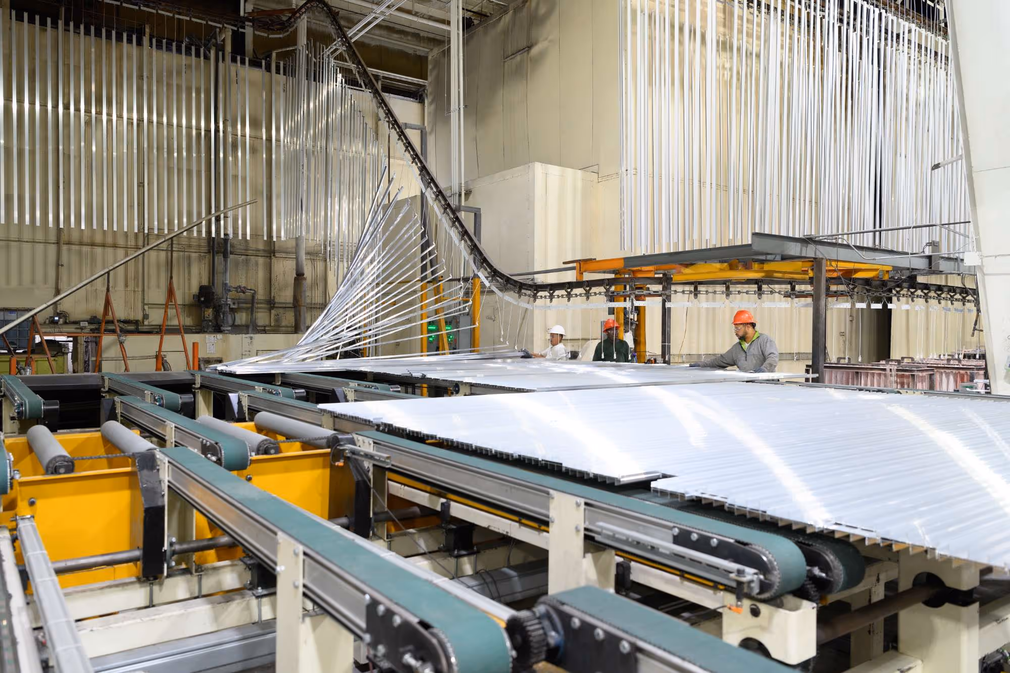 Workers in hard hats inspecting large clear plastic sheets on conveyor belts in an industrial manufacturing facility.