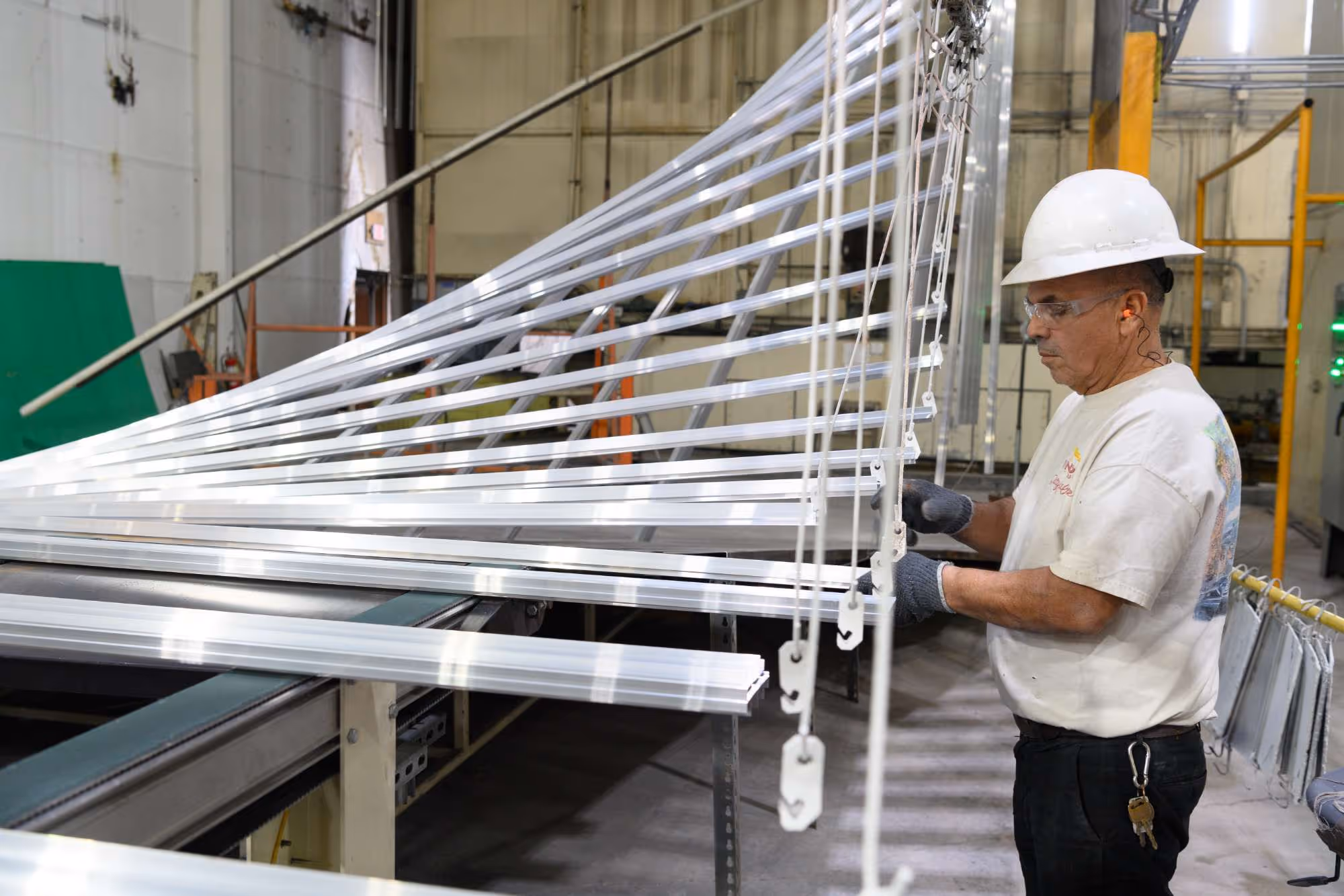 Worker in a white hard hat and gloves inspecting aluminum blinds in a factory setting.