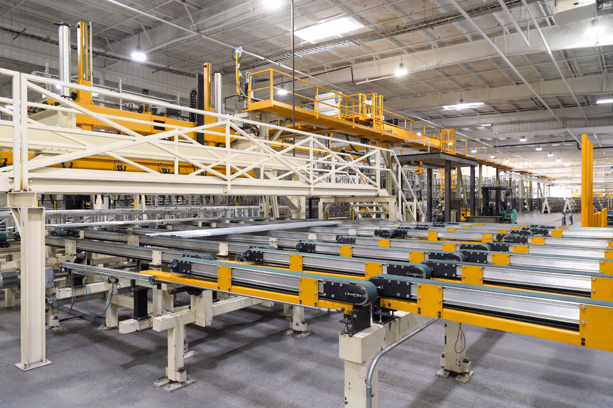 Automated conveyor system with metal rollers in a large industrial warehouse with yellow safety railings and overhead lighting.