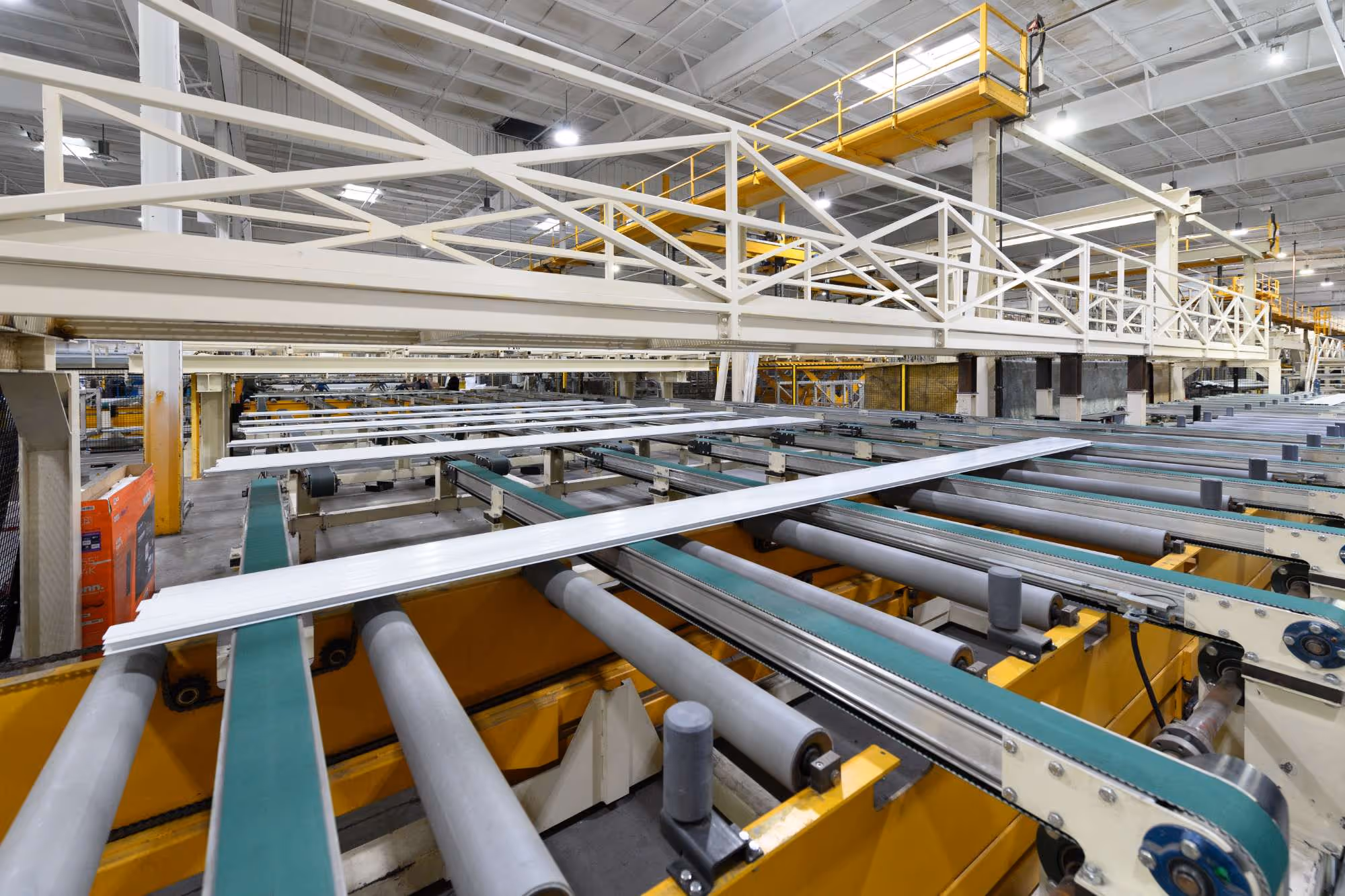 Industrial factory conveyor system with metal rollers and belts transporting flat white panels under a high ceiling with metal support beams.