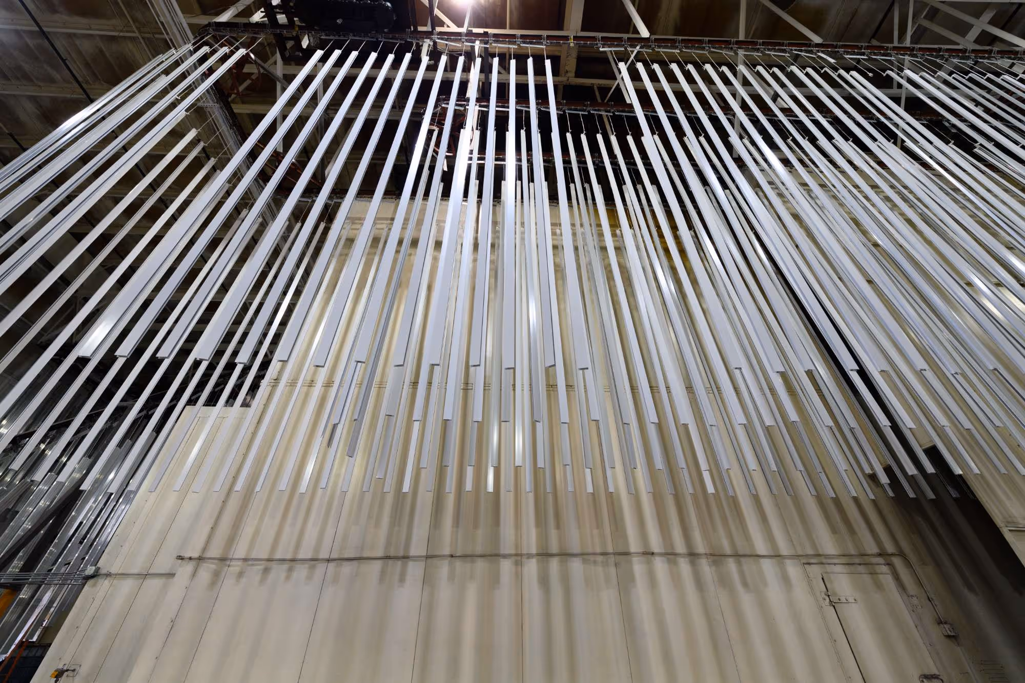 Long metallic bars hanging from a ceiling inside an industrial warehouse casting shadows on a beige wall.