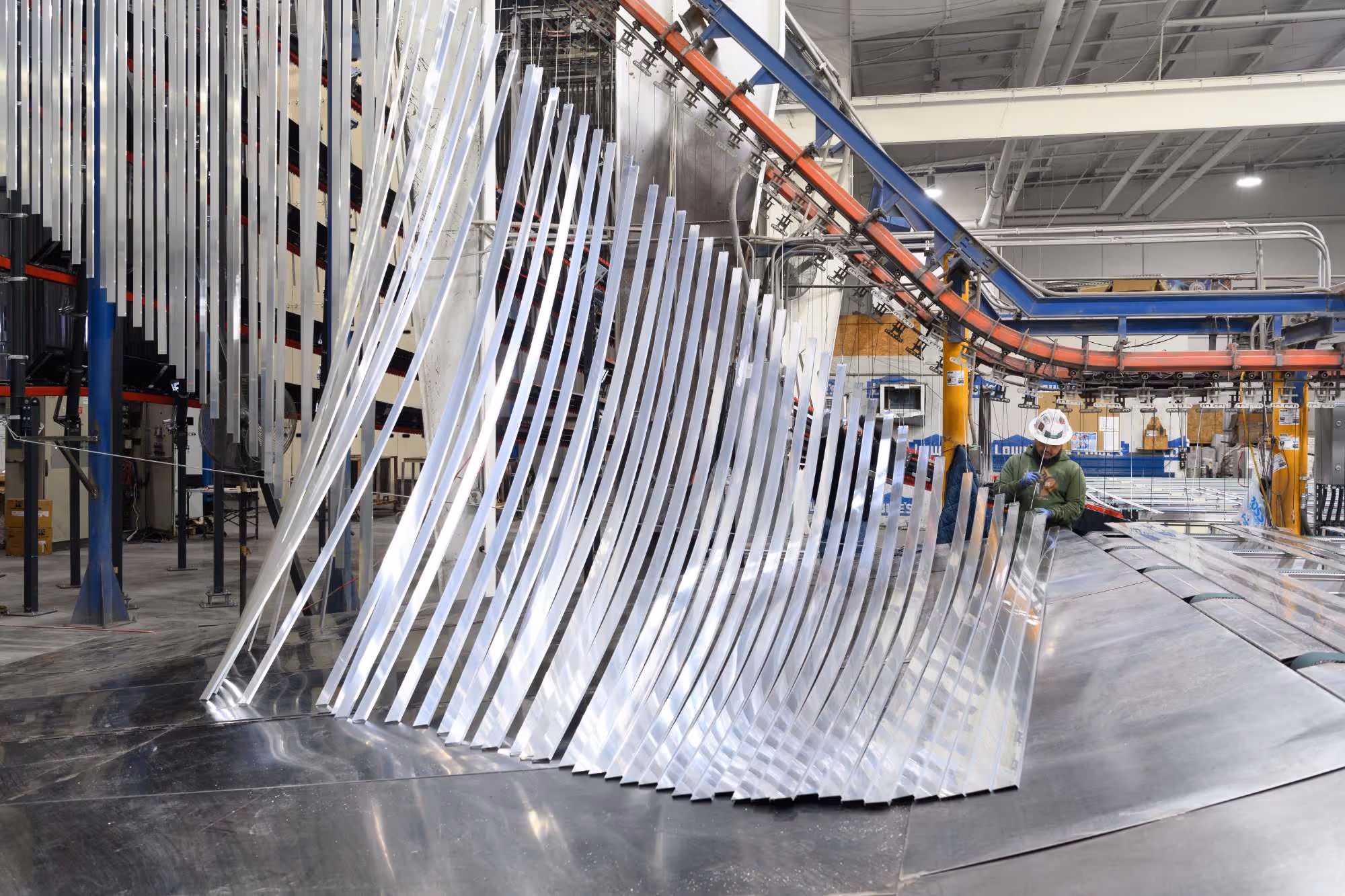Industrial setting with an array of shiny curved metal sheets arranged vertically and a worker wearing a hard hat inspecting one sheet.